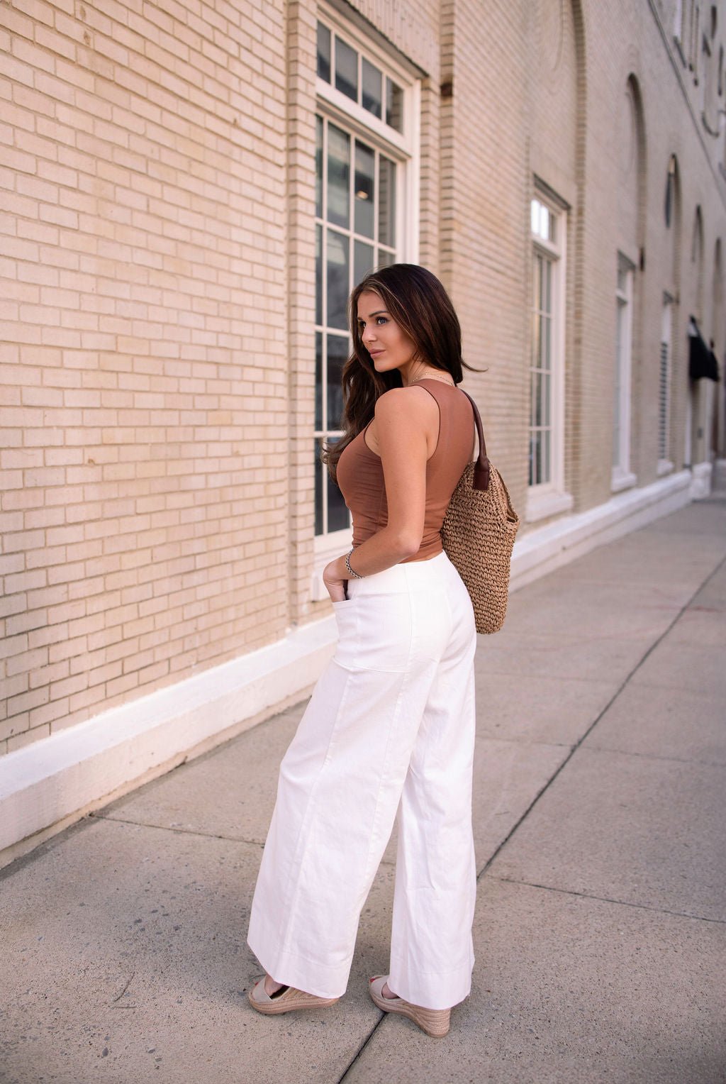 A woman in Deluxe Ivory Wide Leg Denim Pants stands on a sidewalk, looking back over her shoulder with a woven bag.