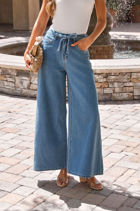 Woman in Denim Belted Wide Leg Pants, a white sleeveless top, and sandals holds a woven clutch by a fountain.