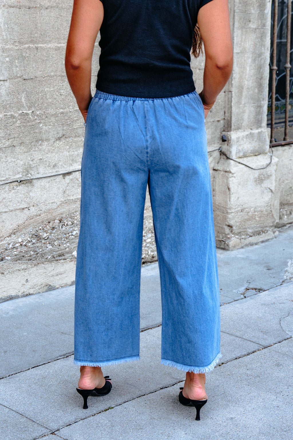 Someone wearing Denim Light Wash Fringe Wide Leg Pants - DOORBUSTER stands on a sidewalk, facing away, in black heeled sandals.