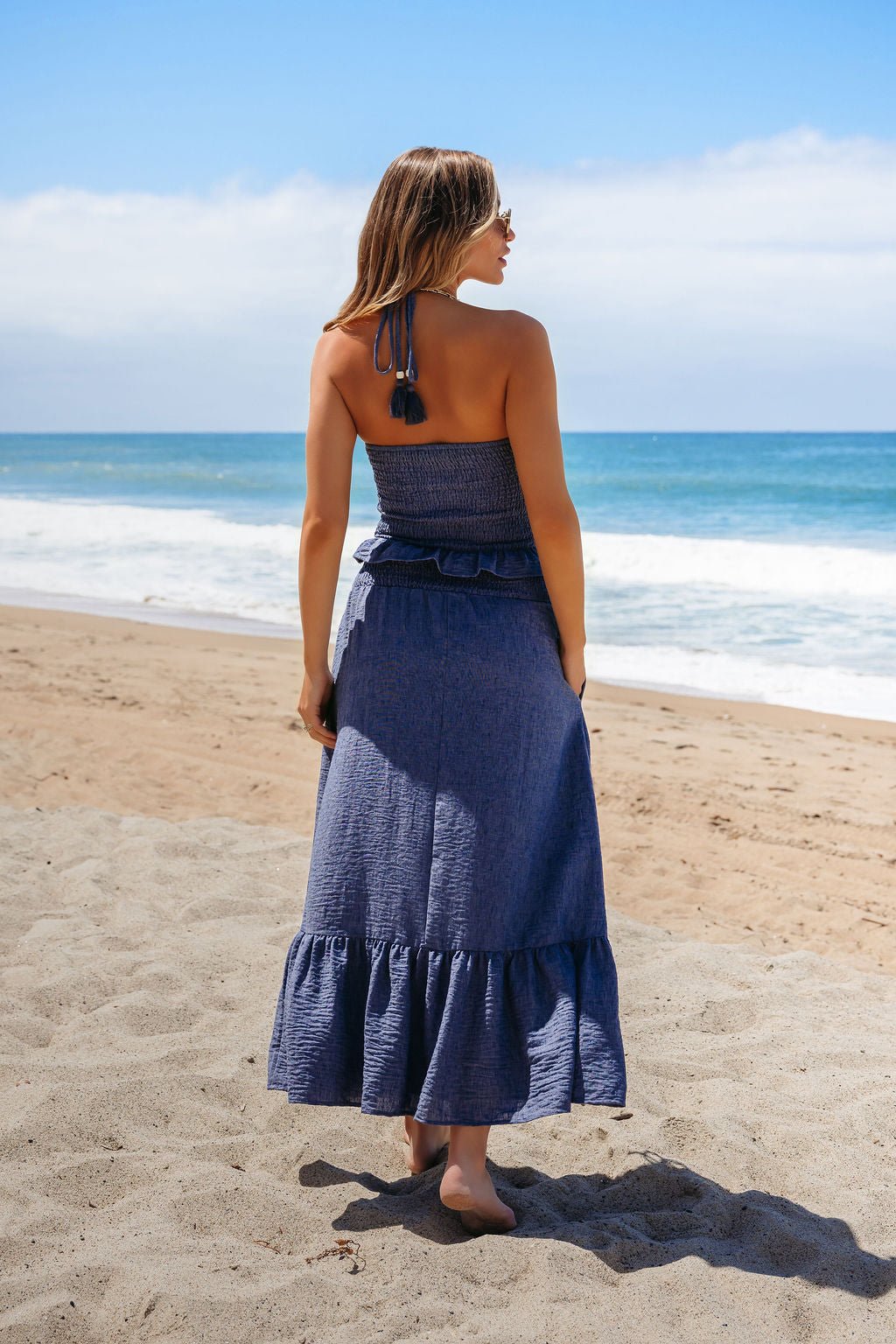 Barefoot woman on a sandy beach faces the ocean in a Denim Smocked Ruffle Halter Tank Top under partly cloudy skies.