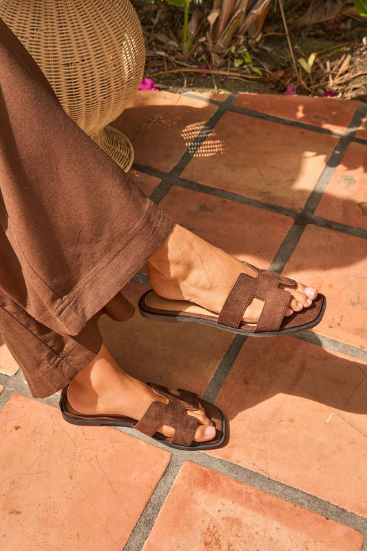 Wearing Desert Drift Sandals - Brown with a suede-look strap and brown pants, standing on sunlit terracotta tiles.
