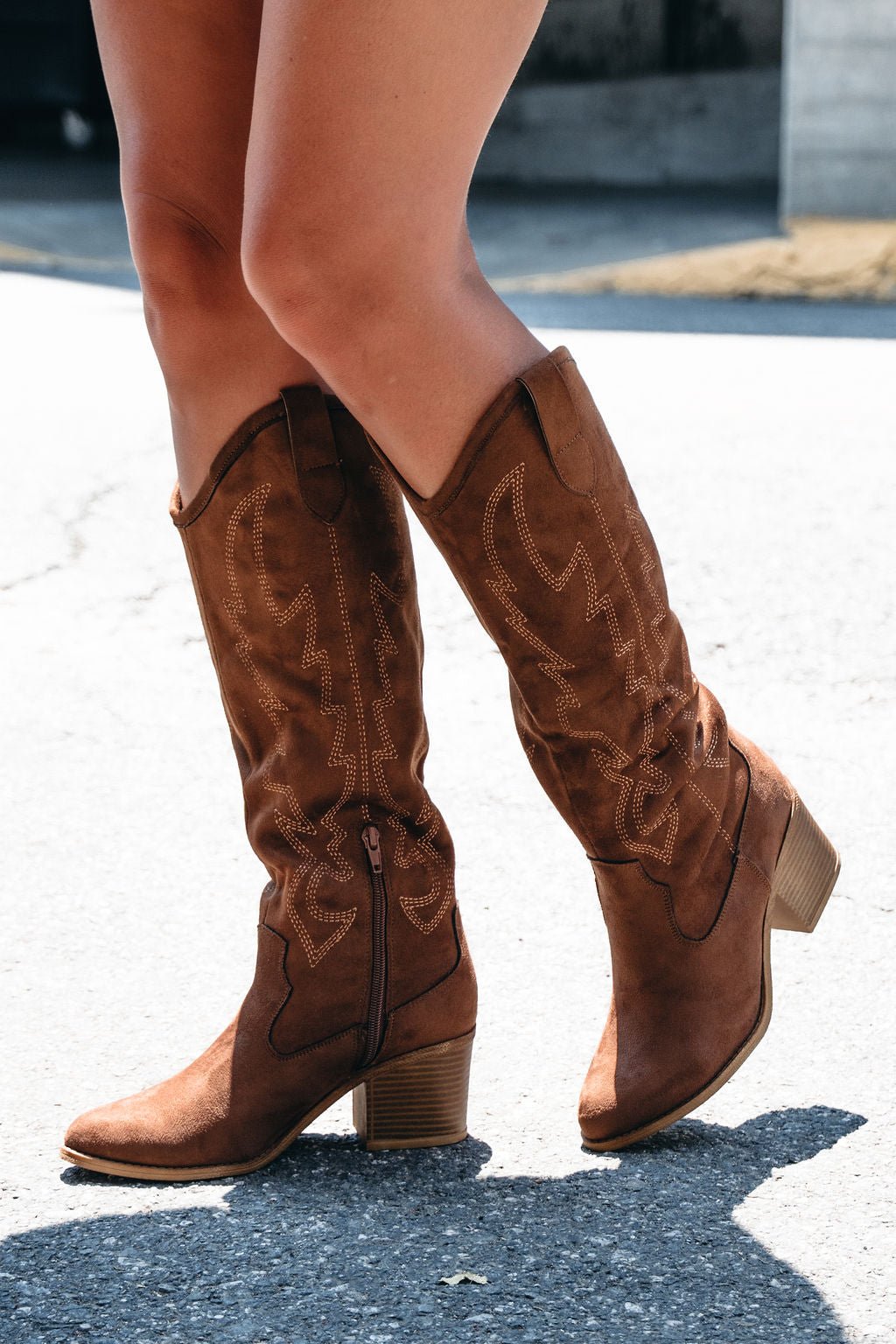 Someone wearing Dirty Laundry Upwind Suede Western Boots in brown stands on a sunlit pavement.
