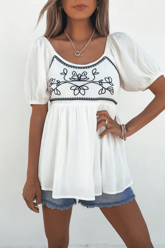 A woman wears the Dream-Filled Moments Ivory Embroidered Babydoll Top with blue shorts, standing against a white background.