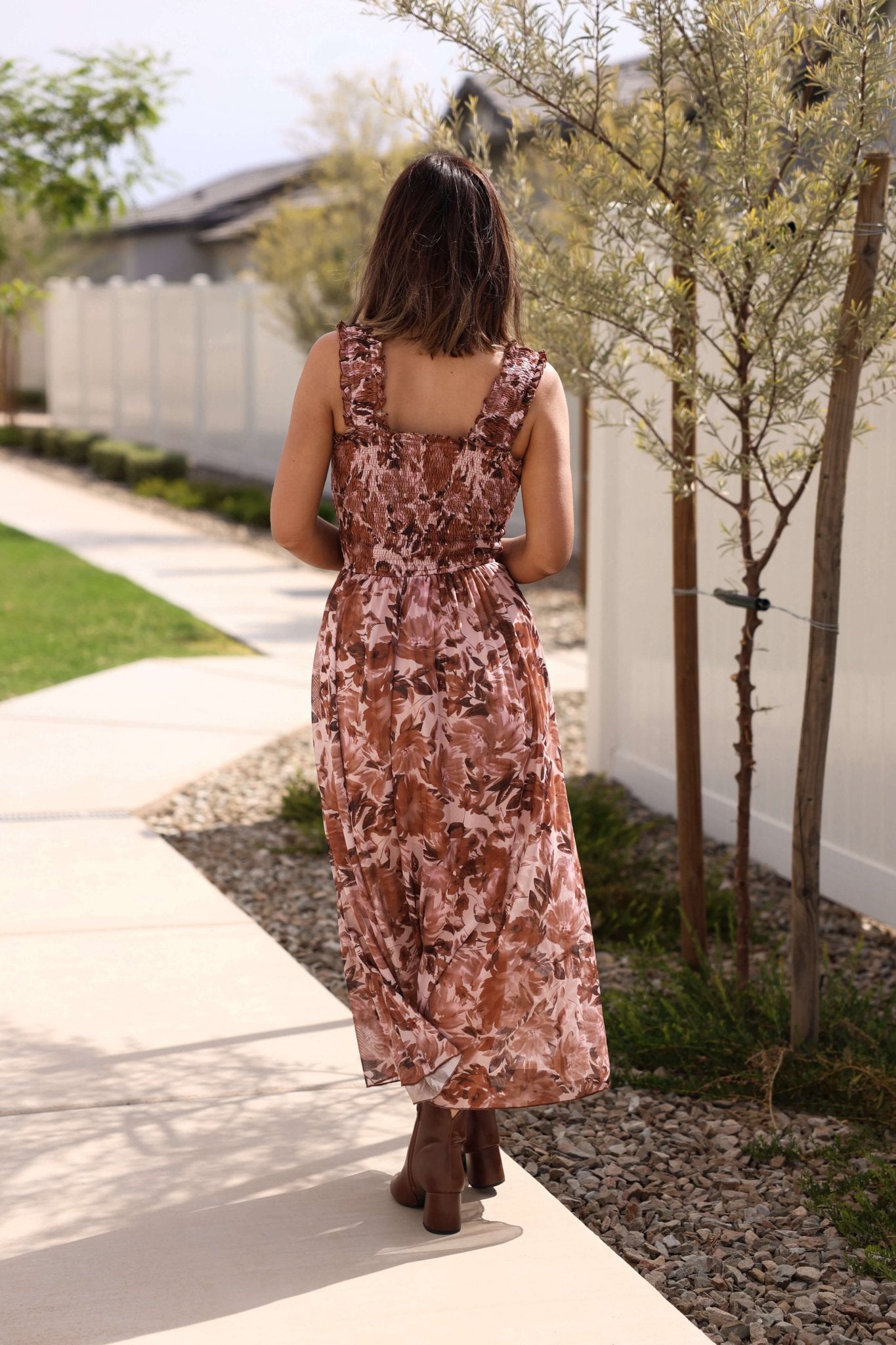 A woman wears the Drifter Brown Floral Print Tiered Maxi Dress, walking on a sidewalk beside a white fence and small trees.