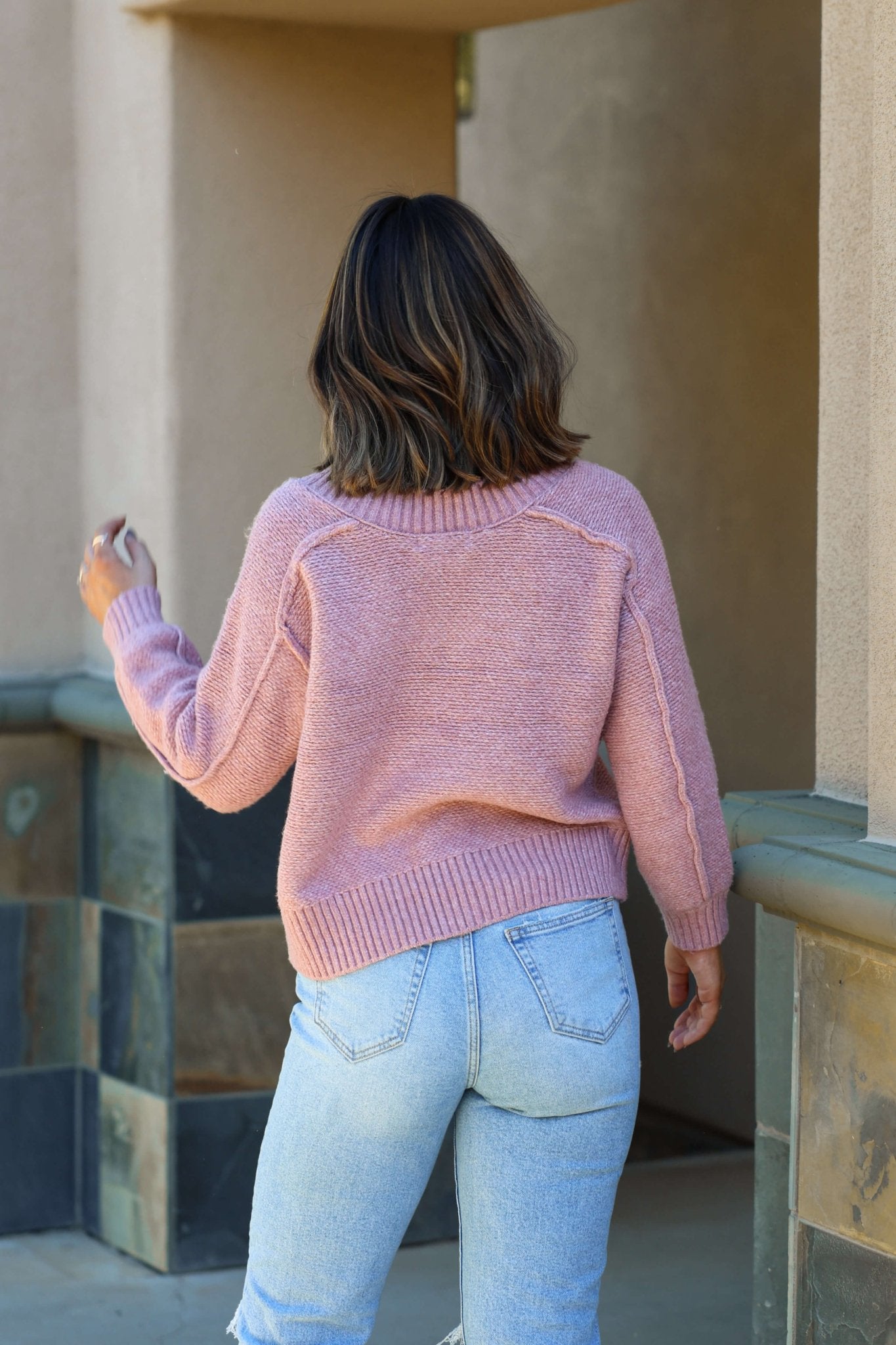 Wearing the Dusty Rose Seam Detail V Neck Sweater and light blue jeans, a person with shoulder-length brown hair walks away.