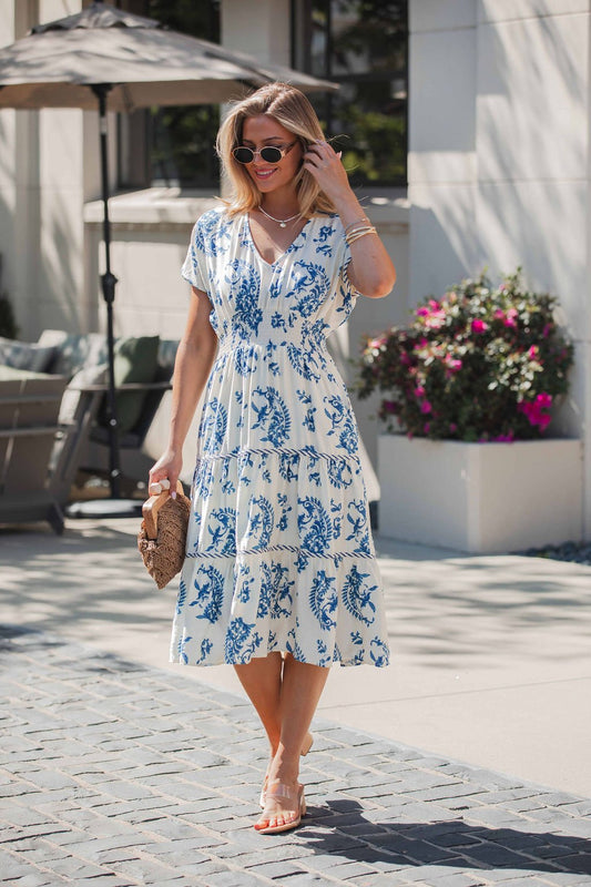 A woman in sunglasses wears the Easton Blue Print Smock Tiered Midi Dress, holding a woven clutch among flowers and patio furniture.