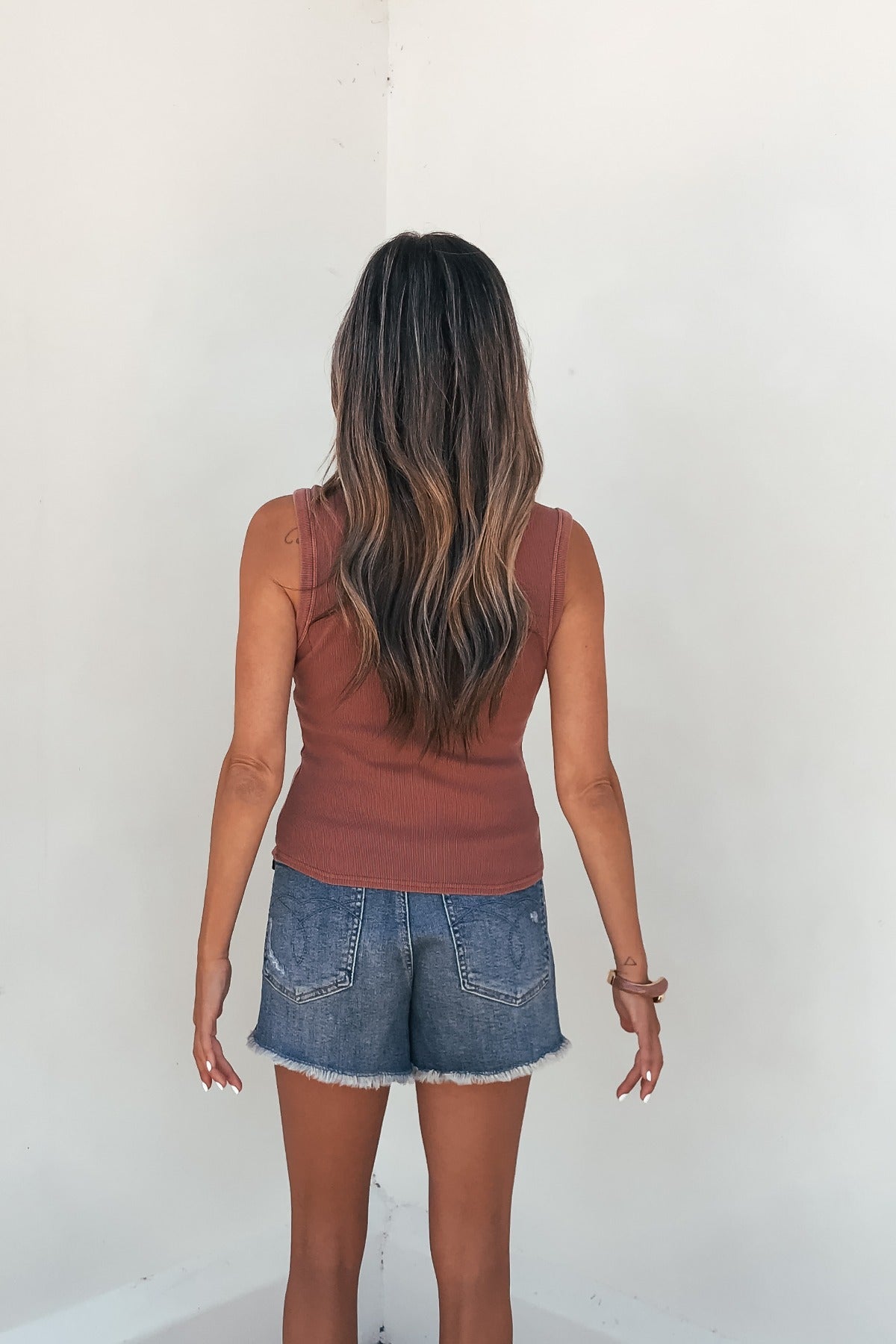 A woman with long wavy hair wears the Easy Days Button Front Tank Top in Terracotta, paired with denim shorts by a white wall.