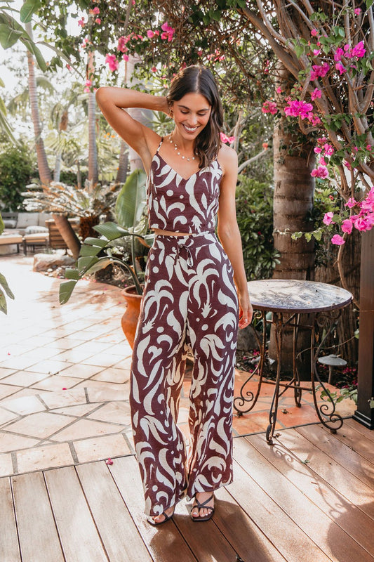 Woman smiles outdoors in the Eda Print Cami Tank and Pants Set - Brown, featuring adjustable cami straps, on a sunny patio.