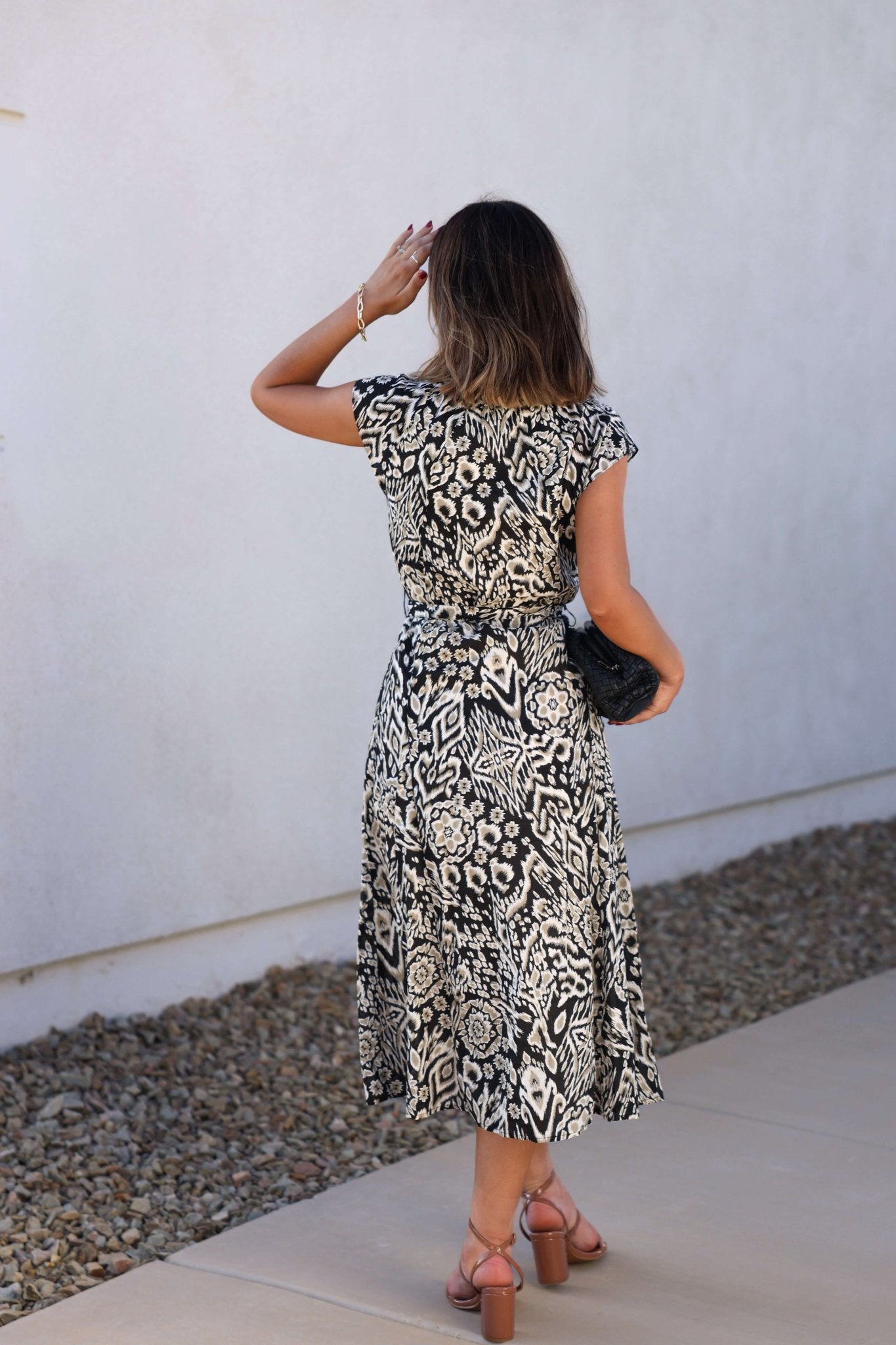 Woman in the Elan Black Print Belted Midi Dress - FINAL SALE stands on a walkway, facing away and touching her hair.