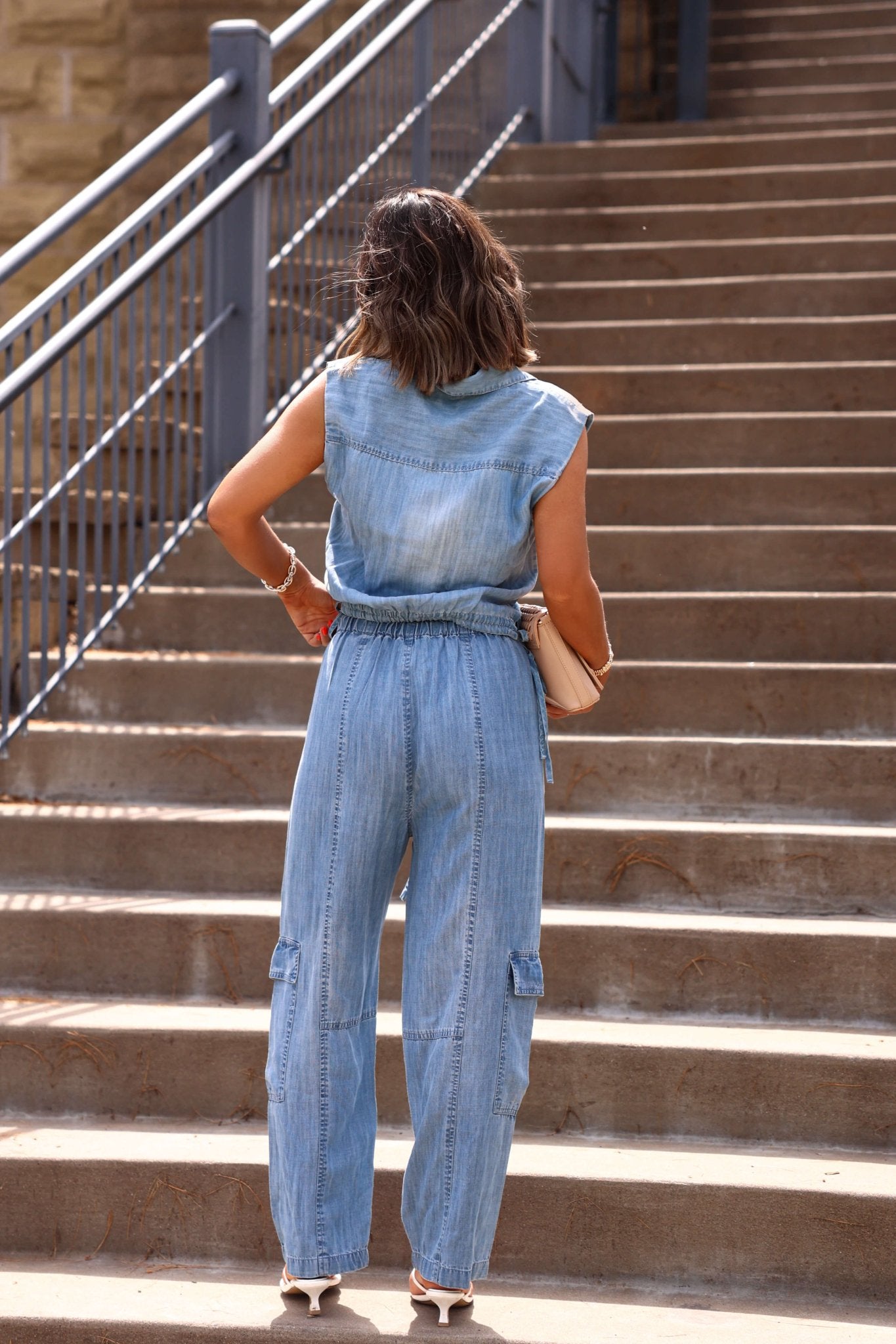 A woman stands at outdoor stairs in Elan Jetset Denim Cargo Pants, facing away and holding a beige purse.