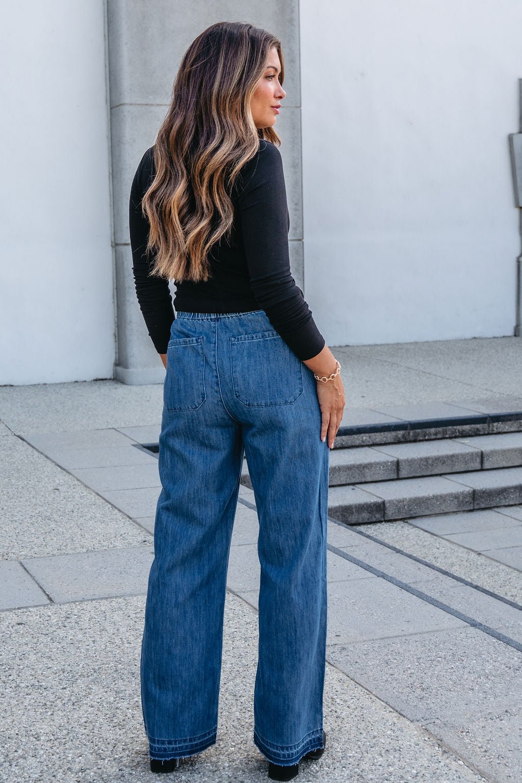 Woman with long wavy hair in a black top and Elan Medium Wash Wide Leg Pants stands outdoors on a stone walkway.