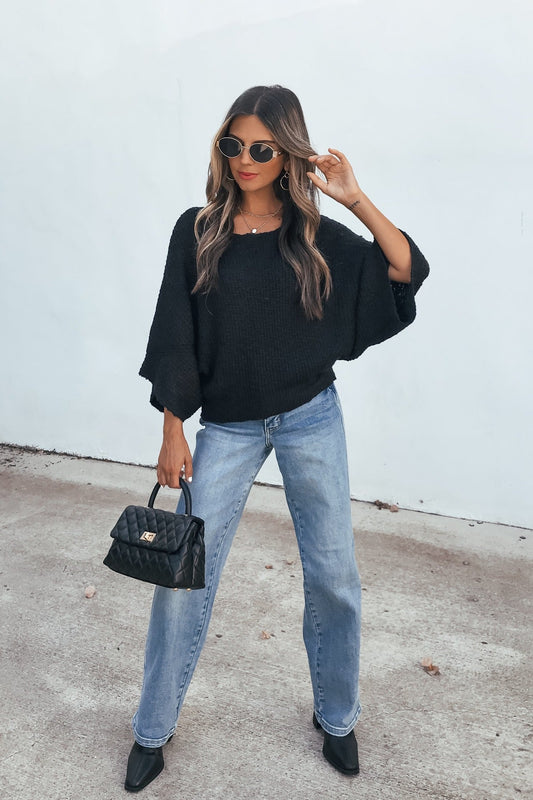 Woman wears sunglasses, jeans, black heels, a purse, and the Elevated Ribbed Boatneck Sweater - Black | Pre Order, against a white wall.