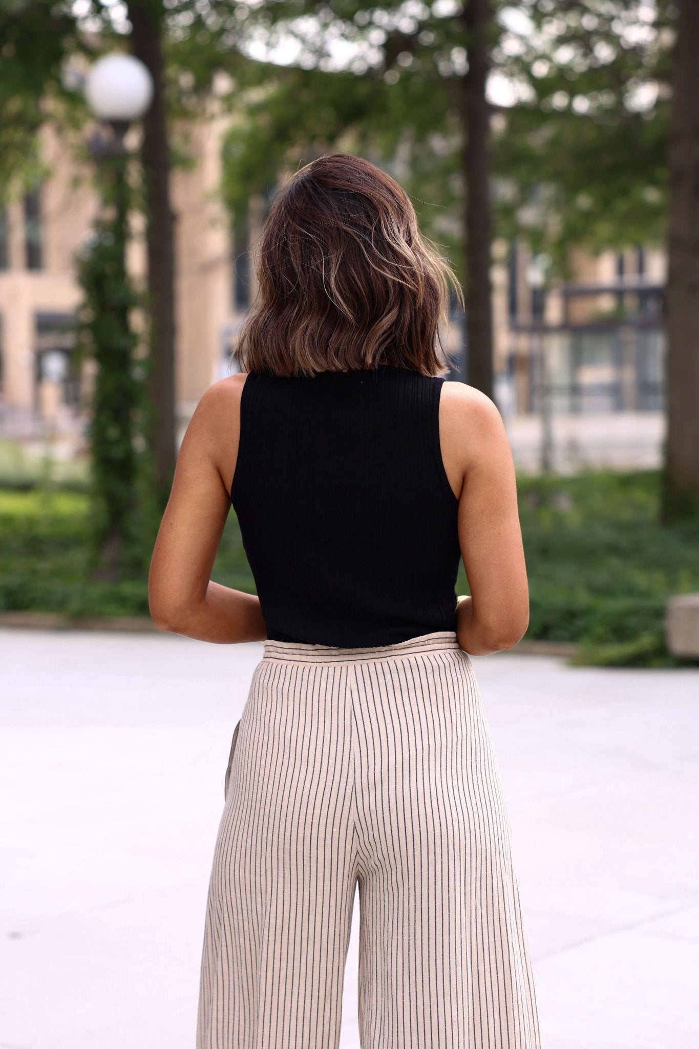 A woman in the Elise Black High Neck Ribbed Tank Top and beige striped pants stands outdoors with her back to the camera.