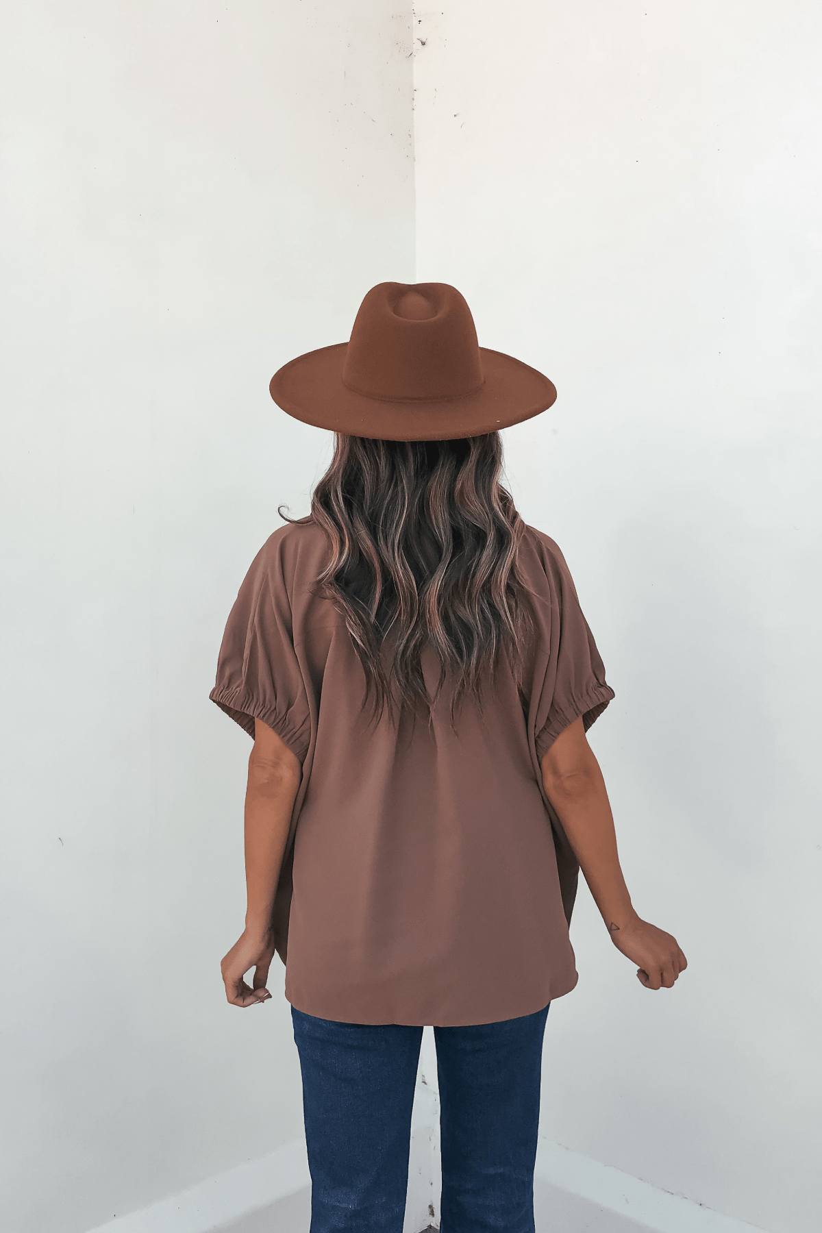A person with wavy hair in a brown hat and the Ellis Camel Split Collared Top stands facing a white corner wall, arms relaxed.