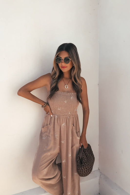 Woman in an Embroidered Smocked Linen Jumpsuit - Latte, with a woven bag, sunglasses, and red lipstick against a white wall.