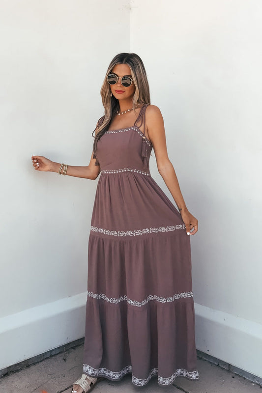 Woman in sunglasses poses against a white wall, wearing the Embroidered Tiered Maxi Sun Dress - Mocha and holding her summer dress.