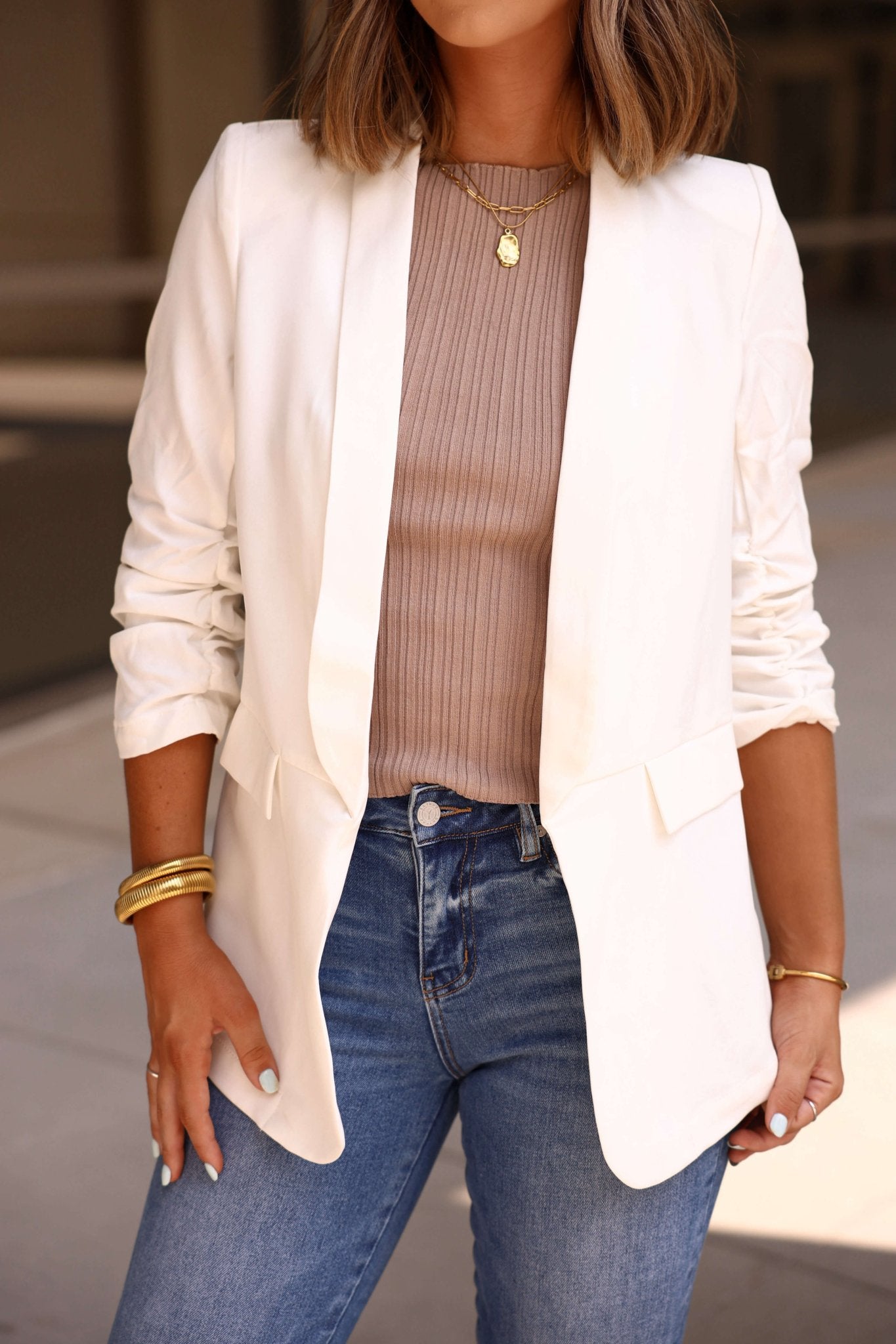 An office-ready look: she's outdoors in a White Ruched Sleeve Blazer - FINAL SALE, taupe ribbed top, blue jeans, and gold jewelry.