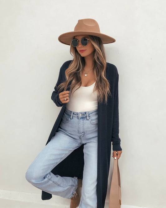 Woman in a tan hat, sunglasses, white top, Essential Navy Duster Cardigan, and light blue jeans poses by a white wall.
