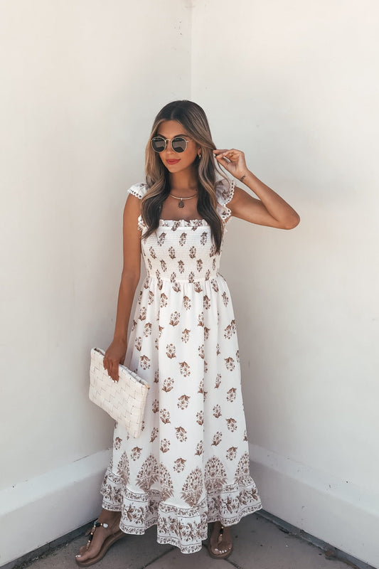 Woman in Everly Smocked Print Midi Dress - Ivory, sunglasses, sandals, and white clutch stands by a plain white wall.