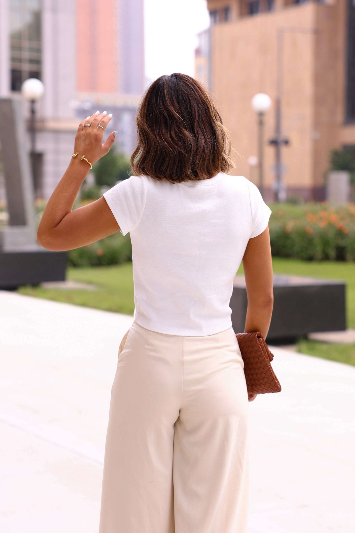Woman with shoulder-length hair waves outside in an Everyday Essential White Tee, beige pants, and a brown clutch.