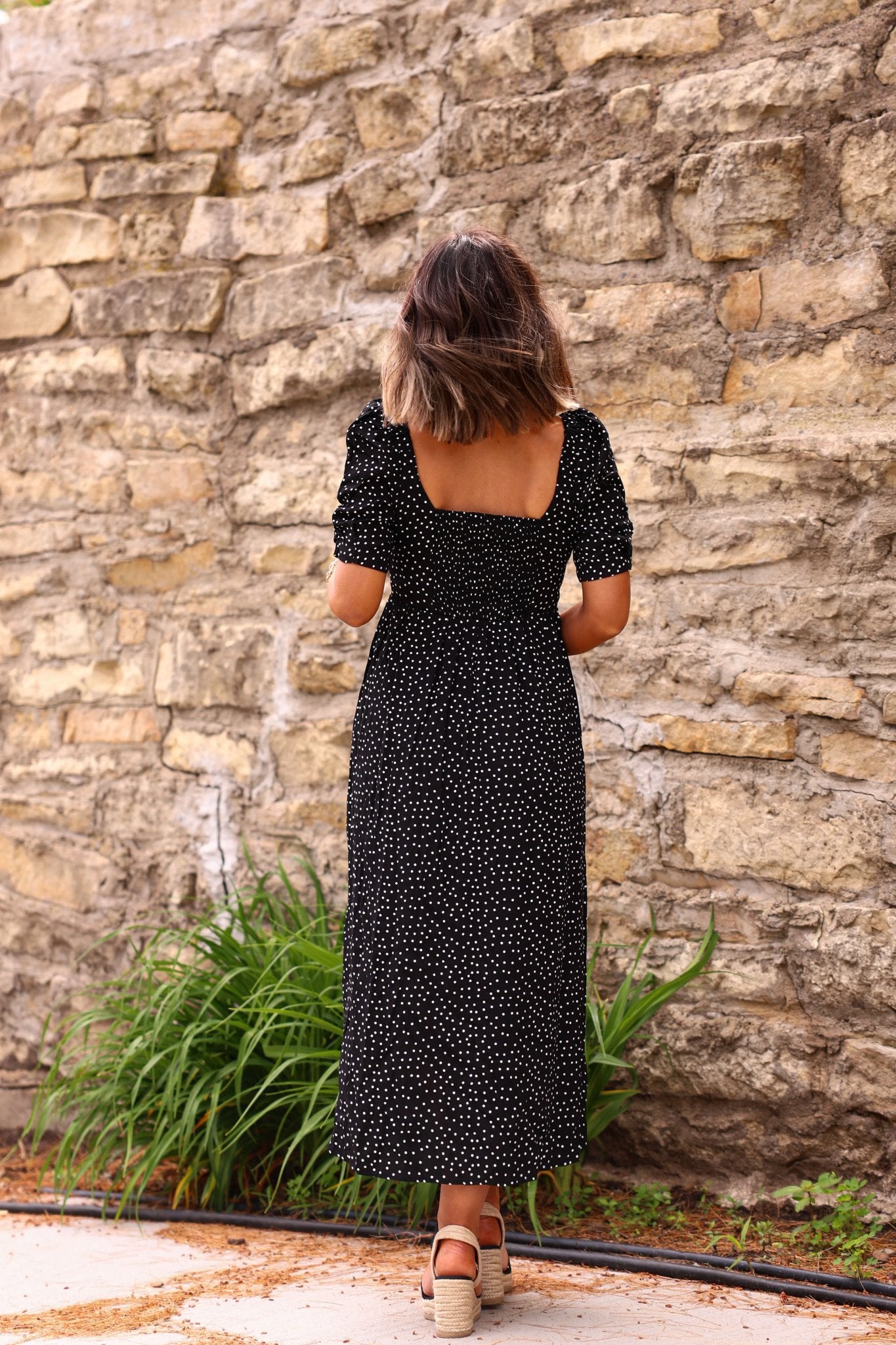 Woman wears the Evie Black Polka Dot Print Midi Dress—FINAL SALE—with sandals, facing a stone wall and green plants.