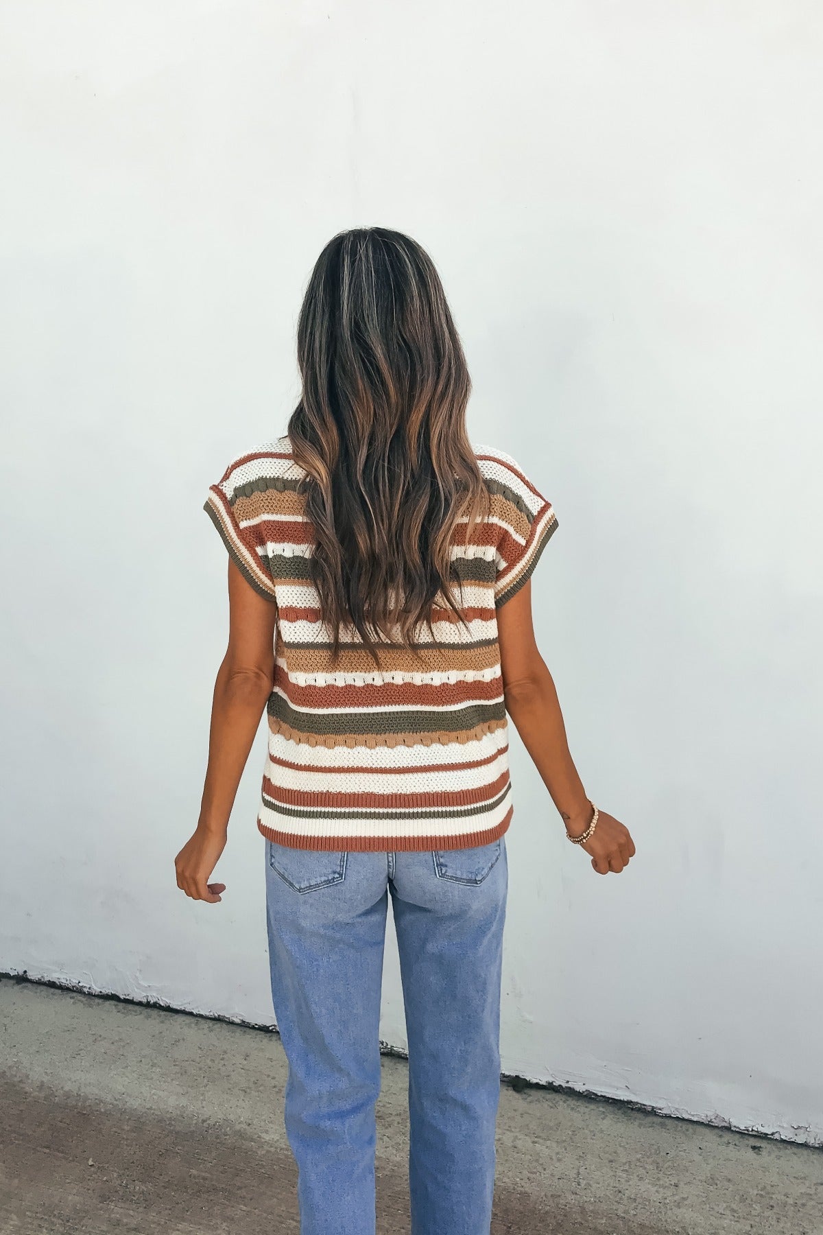 A woman with long wavy hair wears the Field Day Stripe Sweater Vest in olive and blue jeans, standing against a white wall, facing away.