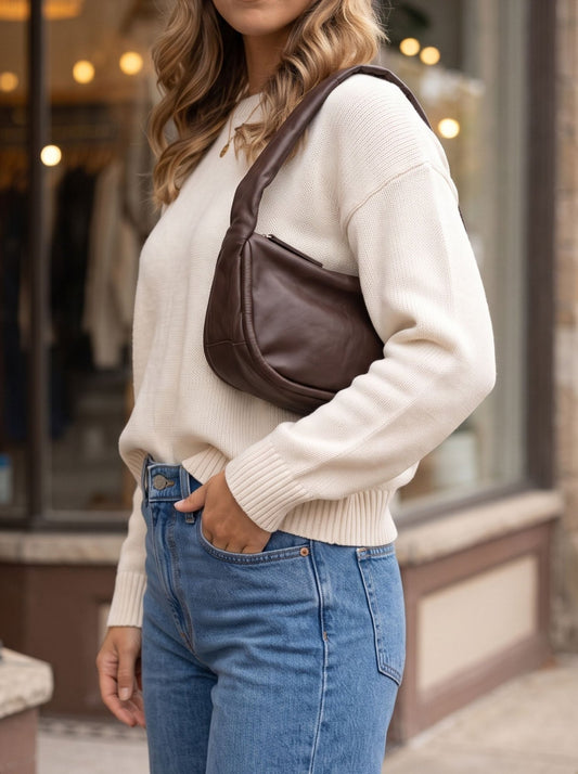 A person in a cream sweater and jeans carries a dark brown, crescent-shaped Free People Alexa Shoulder Bag - Espresso.