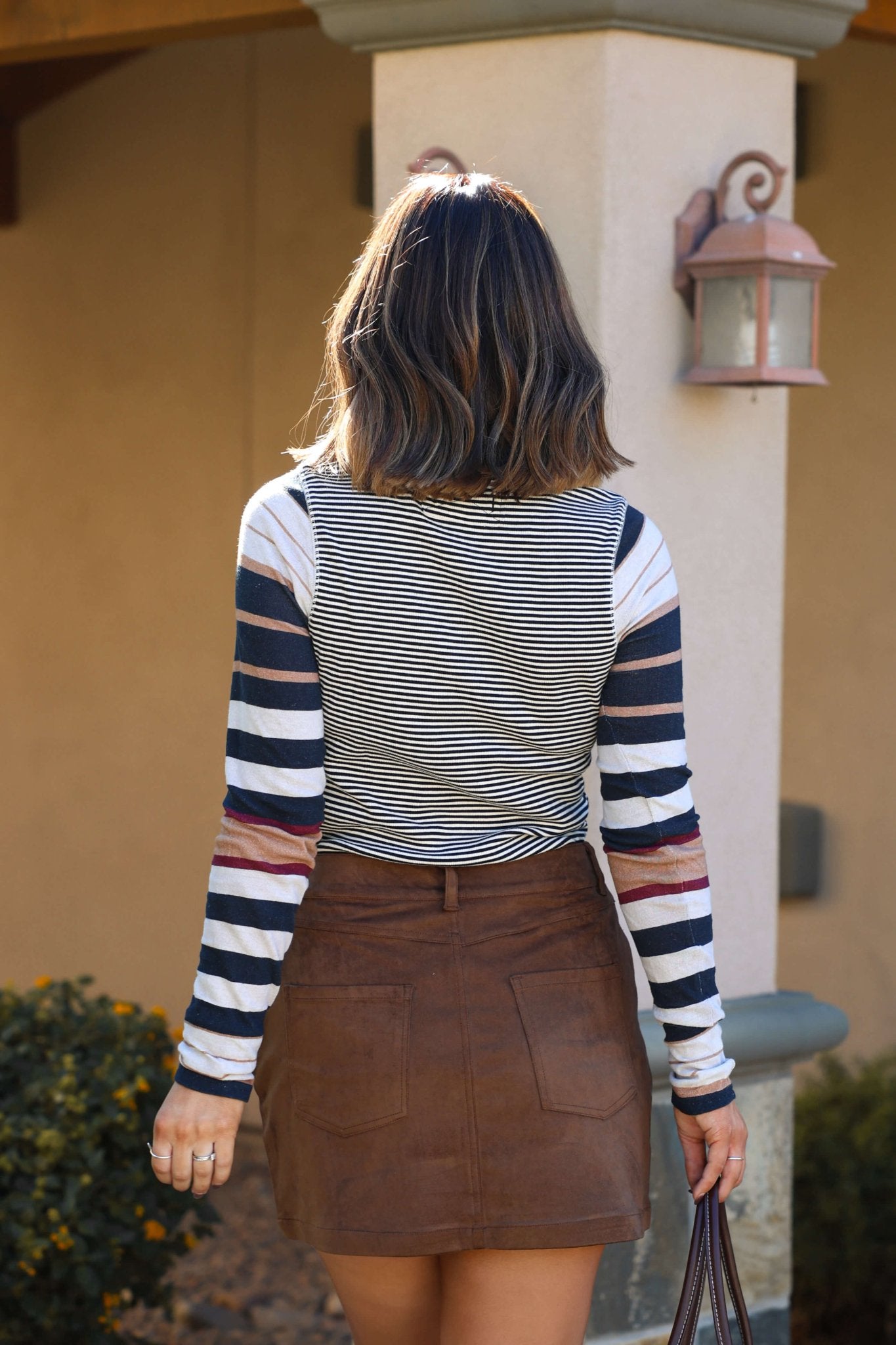 A woman with shoulder-length hair stands outside, facing away, in a Free People Black Combo Avery Zip Top and a brown skirt.