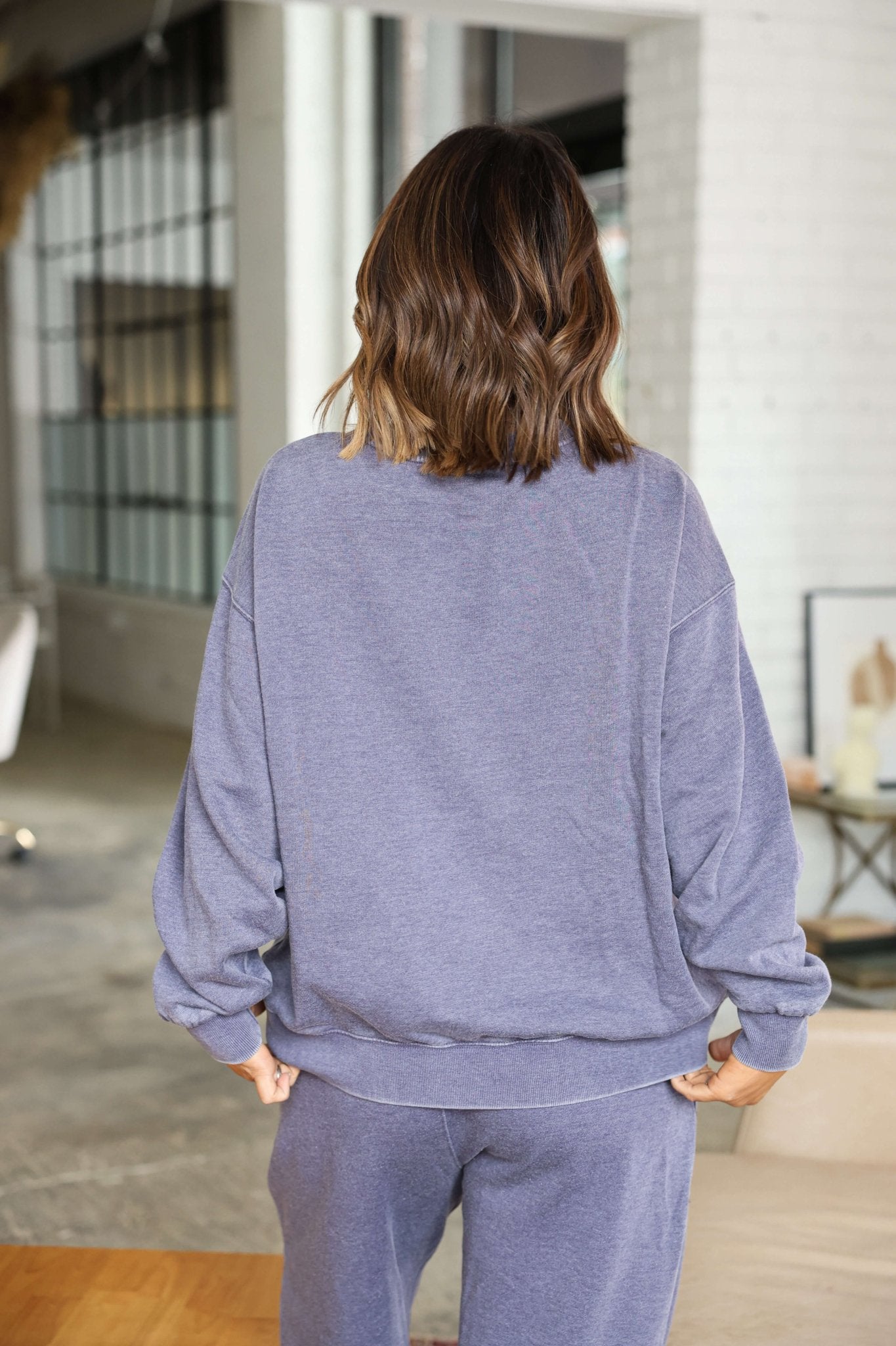 A woman wears the Free People Grey Slow it Down Pullover (FINAL SALE), standing indoors with her back to the camera.