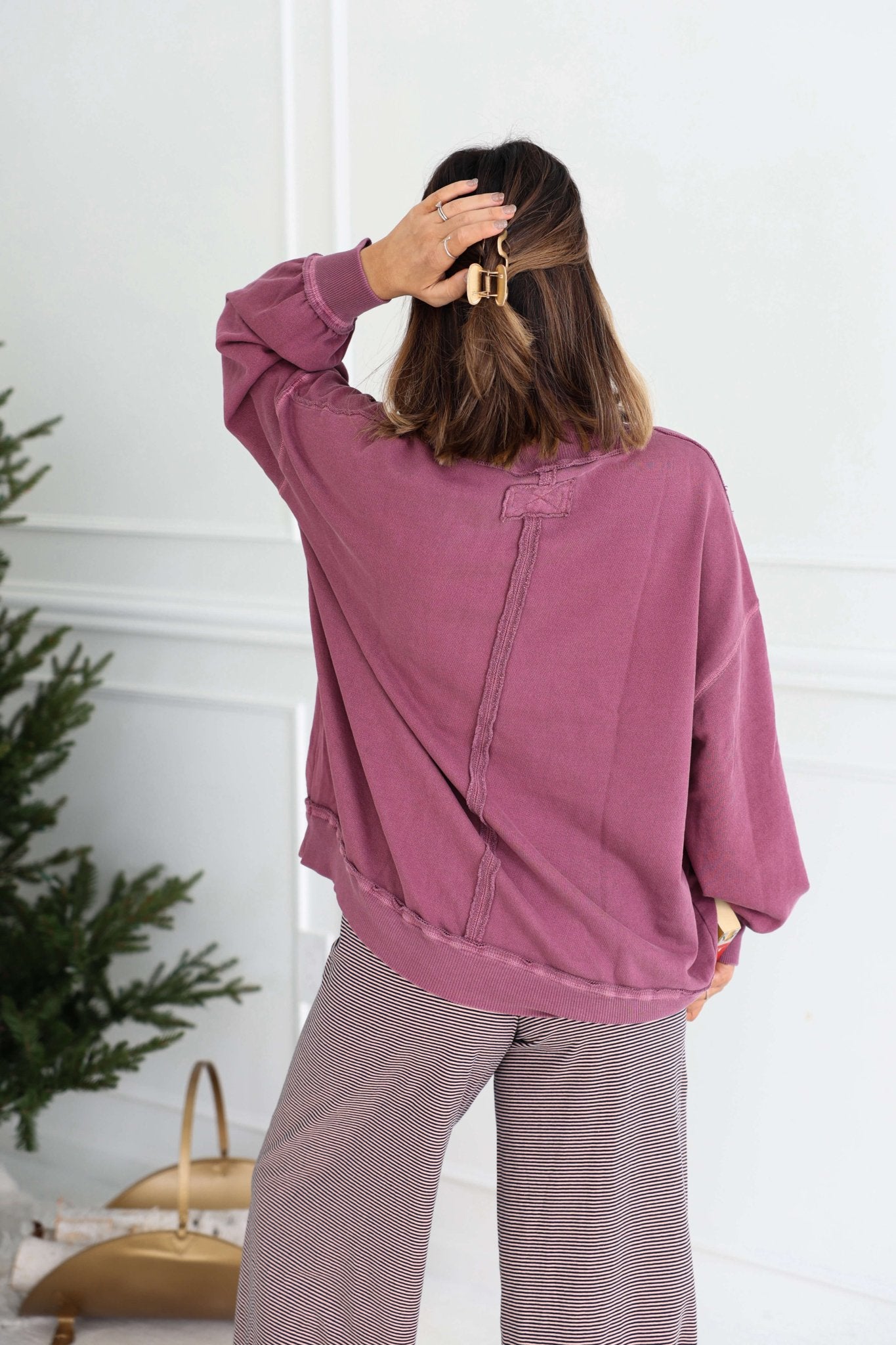 A woman in a Free People Iris Orchid Palm Springs Pullover and gray pants stands with her back to the camera, hand in her hair.