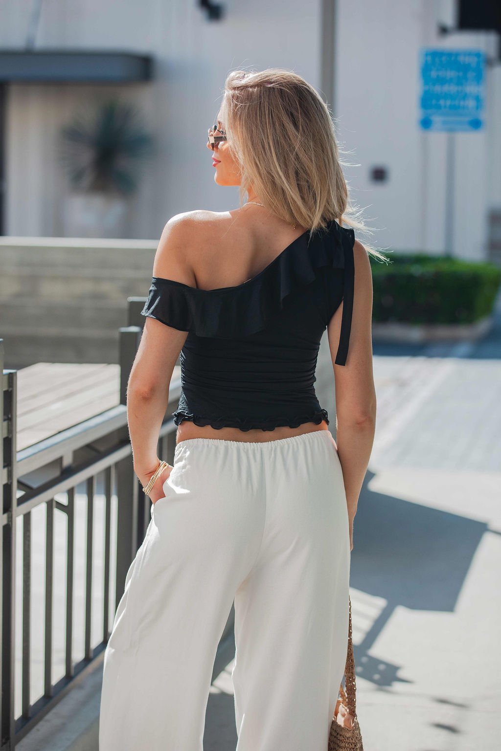 Blonde woman outdoors, facing away, wears a black Free People Isla Off The Shoulder Top and white pants on a sunny day.