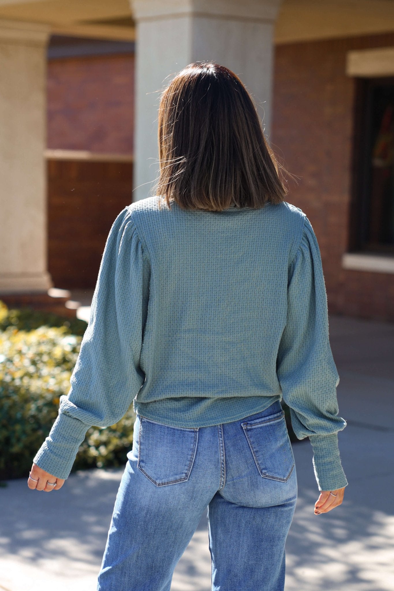 A woman with shoulder-length brown hair wears a Free People Jade Quinn Thermal Top - FINAL SALE and jeans, back to the camera outside.
