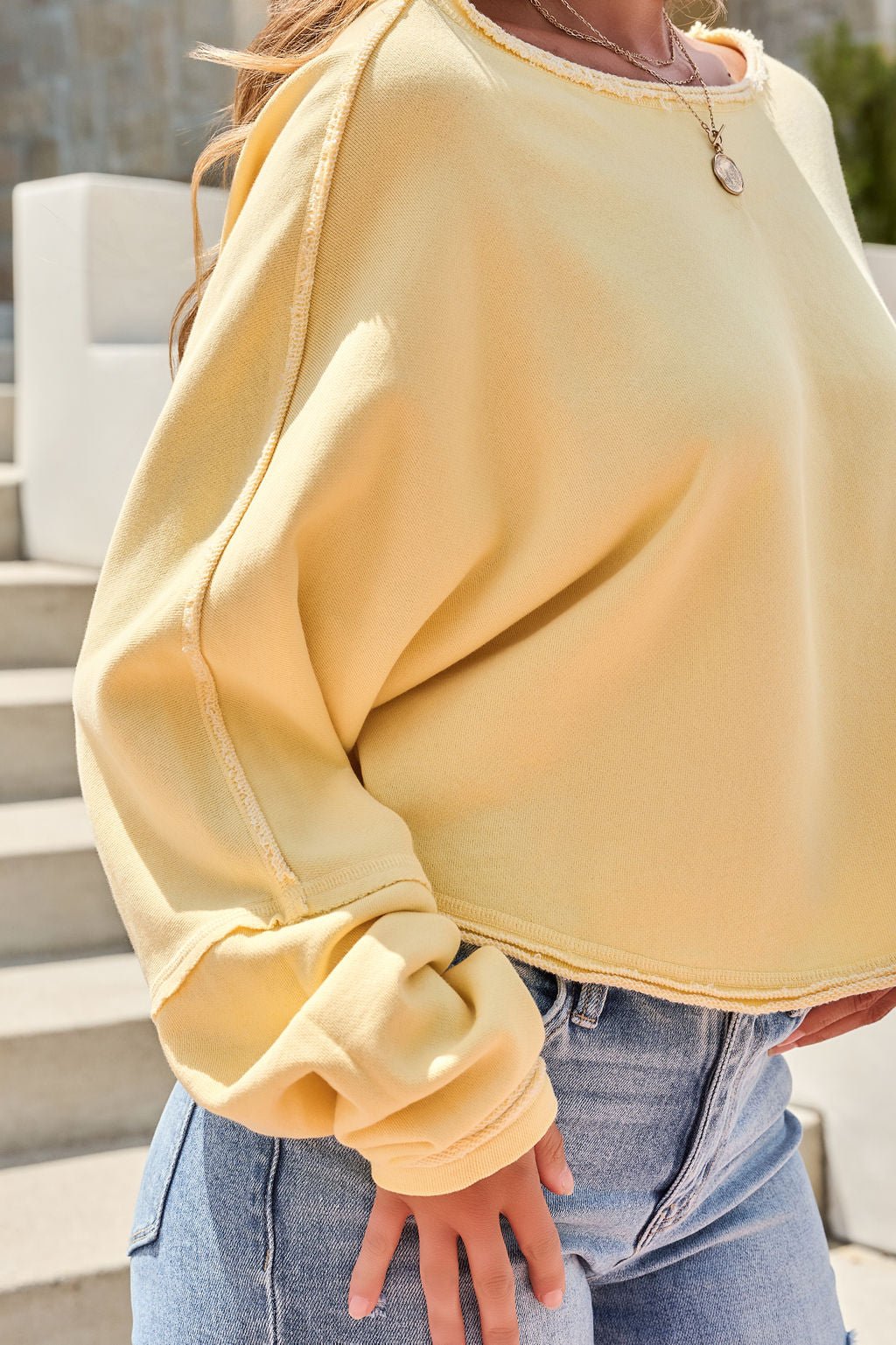 Wearing the Free People To The East Pullover in butter yellow and light blue jeans, a person stands on steps enjoying the sunlight.
