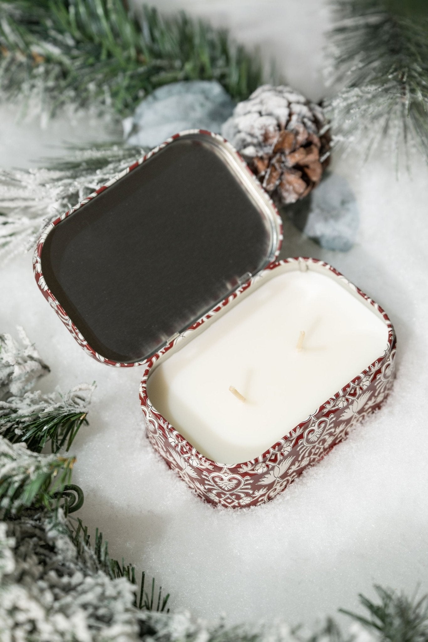 The French Linen & Orris Print Tin Soy Candle comes in a decorative tin with frosted pine, snow, and pinecones.