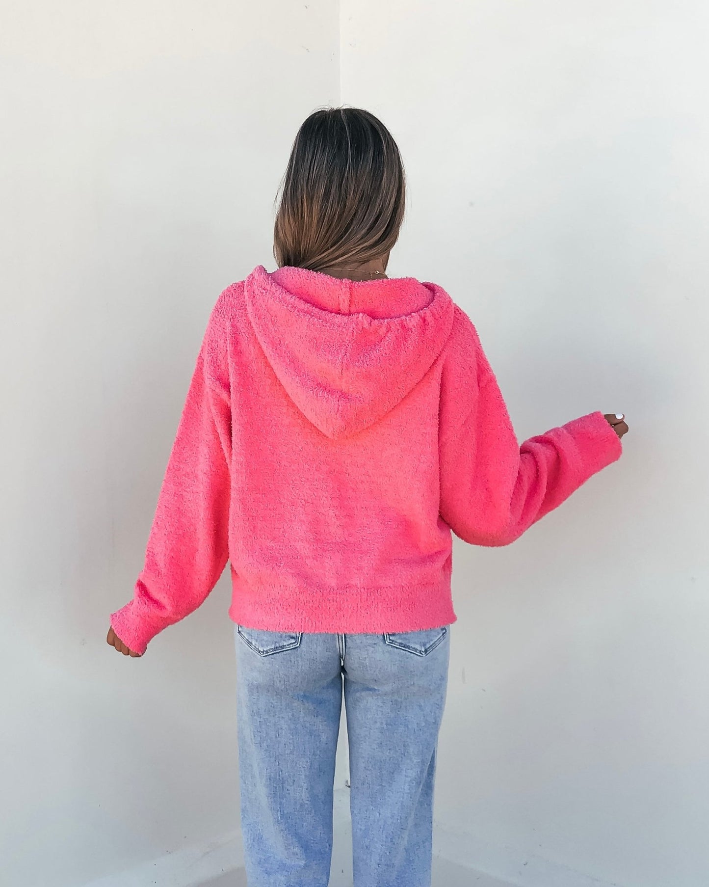 Facing a white wall, a person wears the Fuzzy Pink Hooded Sweater with light blue jeans, arms slightly raised.