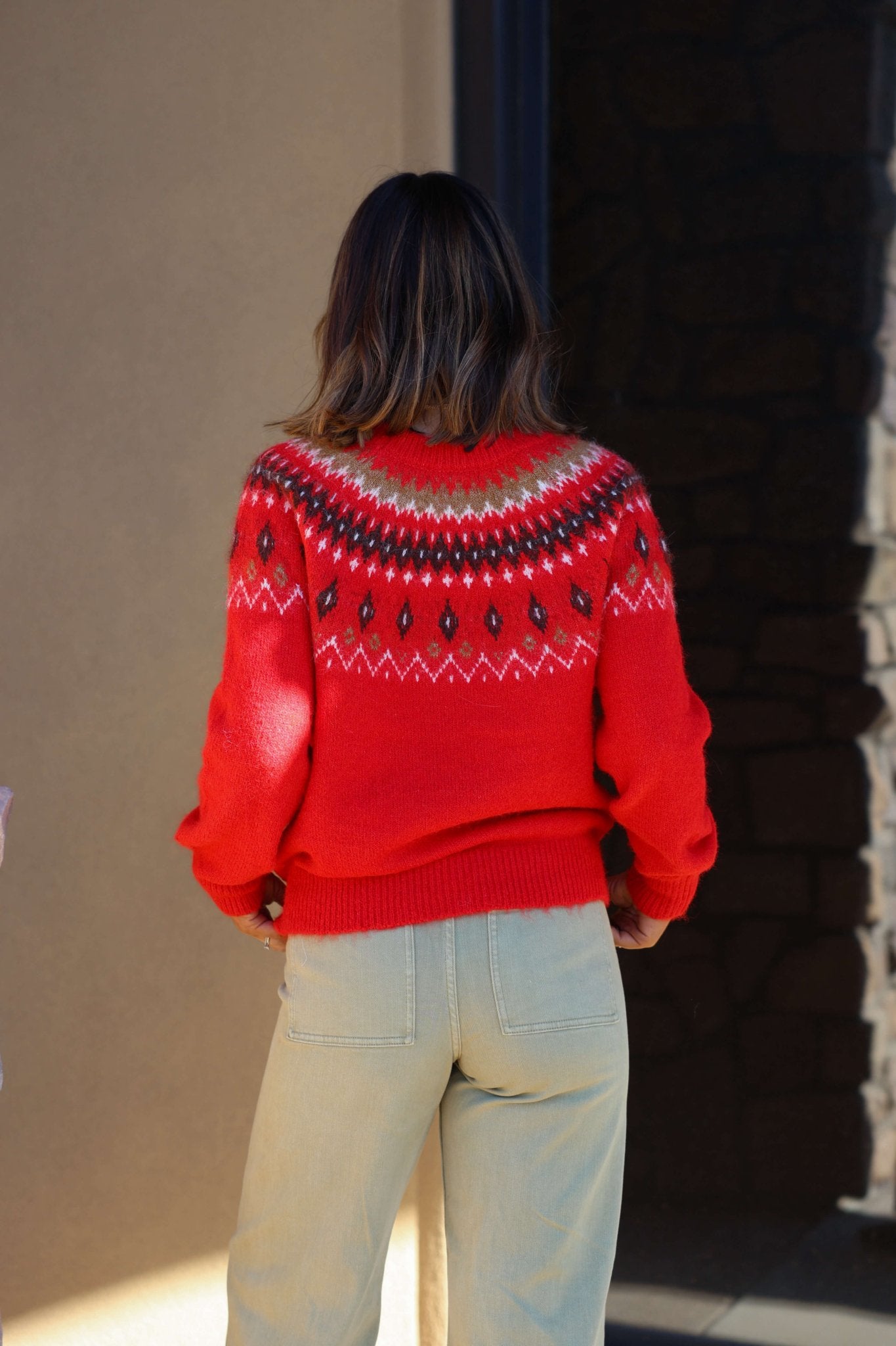 A person with dark hair, seen from behind, wears a Fuzzy Red Fair Isle Crew Neck Sweater and light pants near a dark doorway.