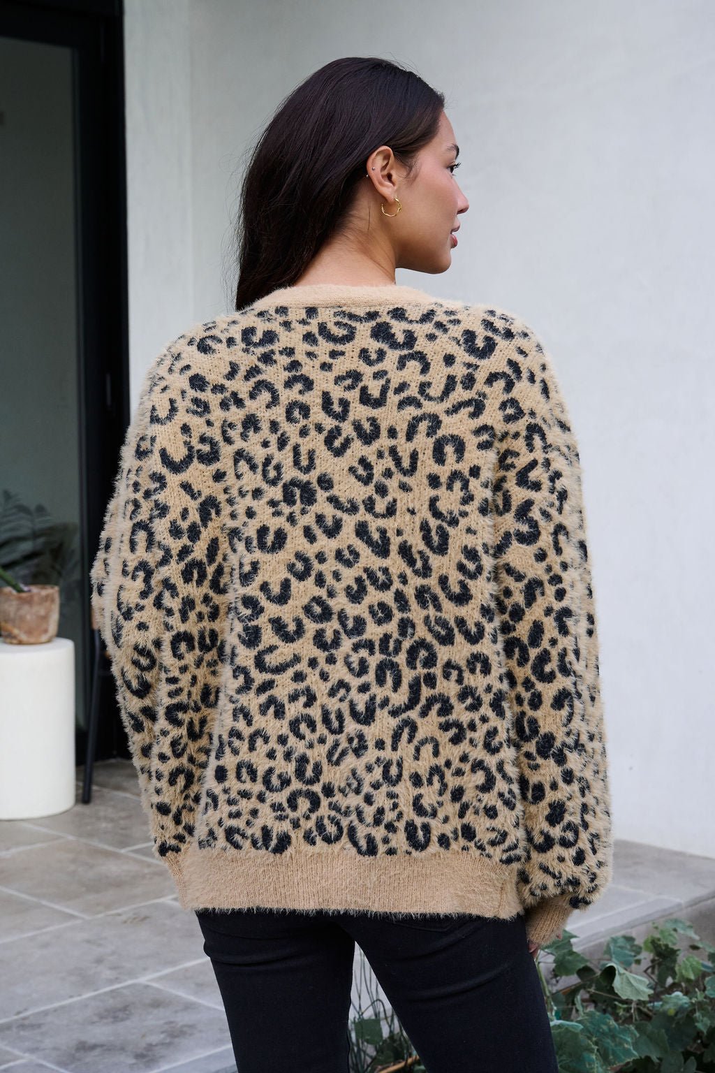 A woman with long dark hair wears the Fuzzy Tan Leopard Open Front Cardigan, standing outside and facing away from the camera.