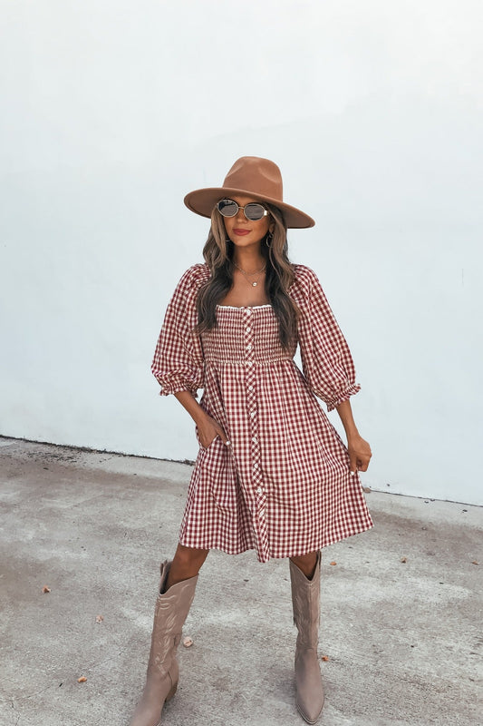 Woman in a brown hat and sunglasses wears the Gingham Babydoll Mini Dress - Burgundy, posing outdoors against a plain white wall.