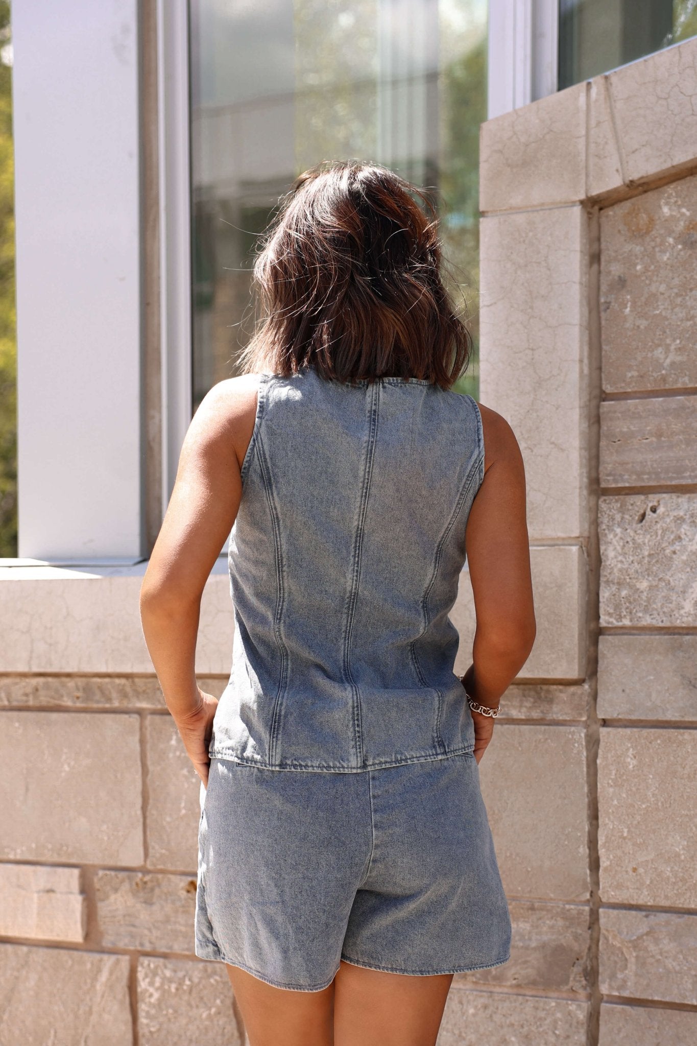 A person with shoulder-length hair wears the Gold Button Down Denim Vest-FINAL SALE while facing a stone wall outdoors.