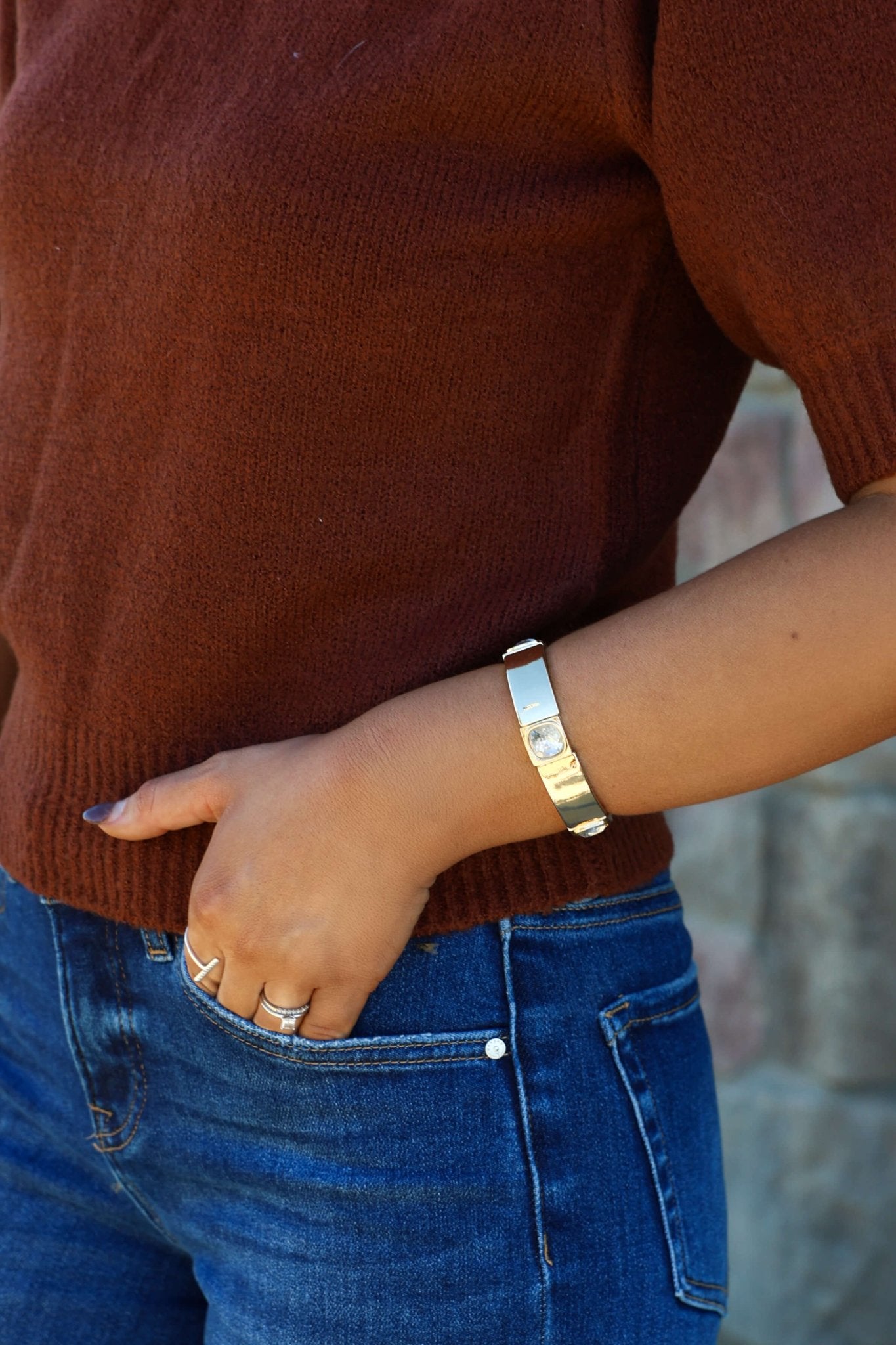 A person in a rust sweater, blue jeans, silver rings, and a Gold Center Gemstone Bracelet stands with one hand in their pocket.