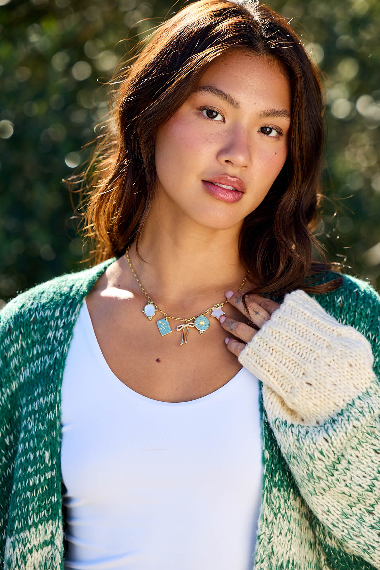 A woman with long brown hair wears a green sweater and the Gold Christie Multi Charm Chain Link Necklace, standing outdoors.