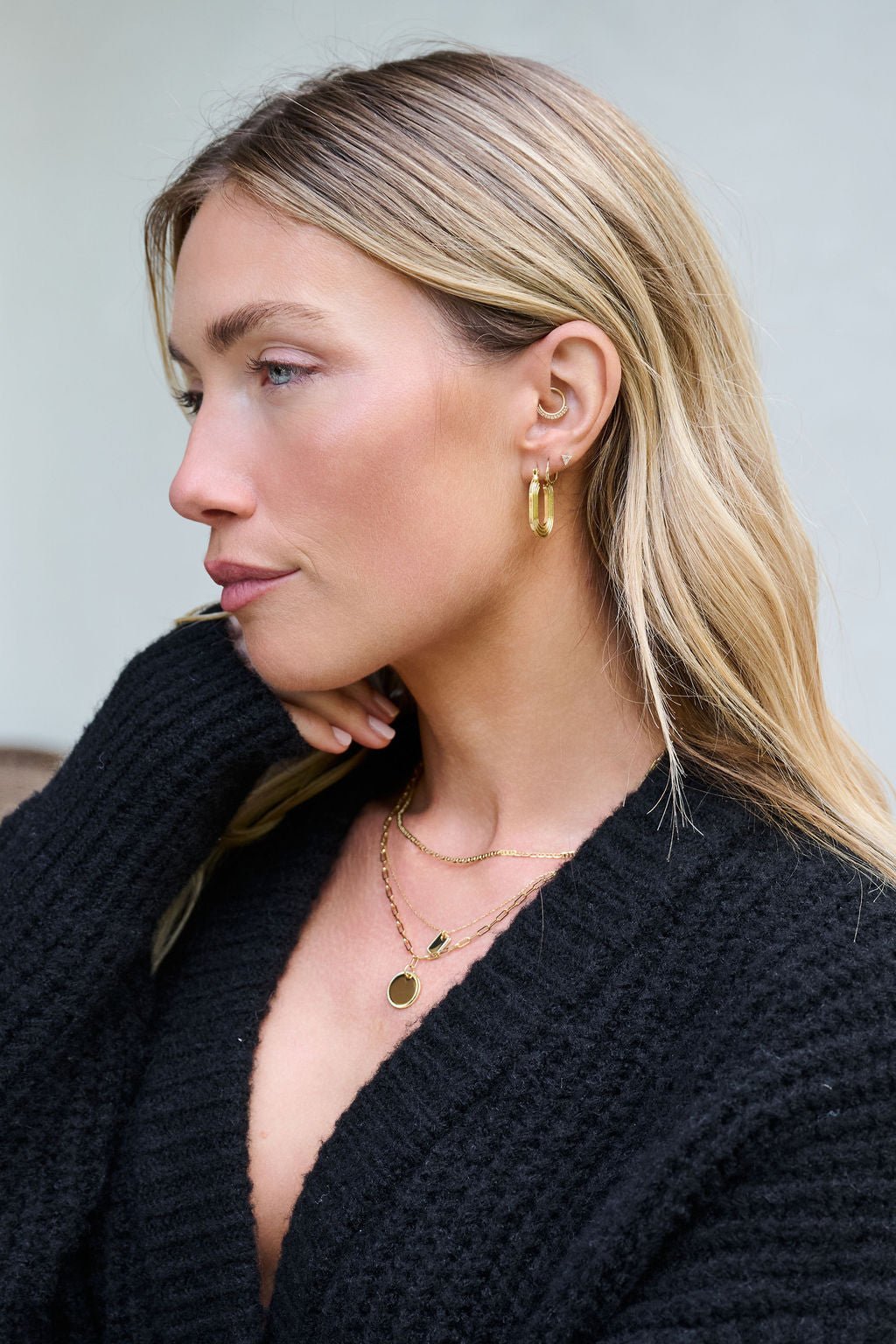 Woman with long blonde hair wears Gold Dipped Flat Grooved Hoop Earrings and a black textured sweater, looking to the side.