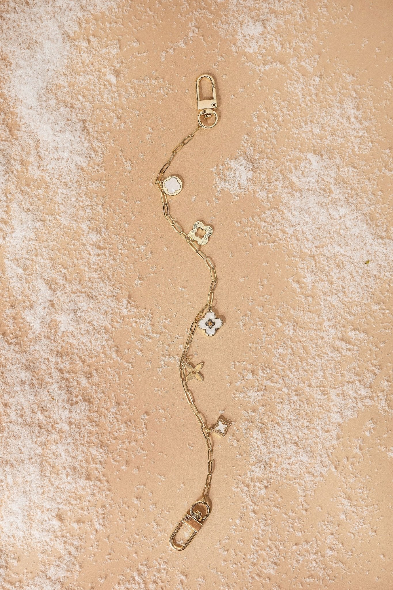 The Gold Multi Clover Charm Purse Chain with floral details rests on a peach surface, perfect as a handbag accessory.