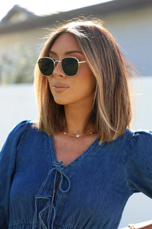 Woman with shoulder-length blonde hair wearing Gold Smoke Lens Narrow Sunglasses, a denim top, and gold necklace outdoors.