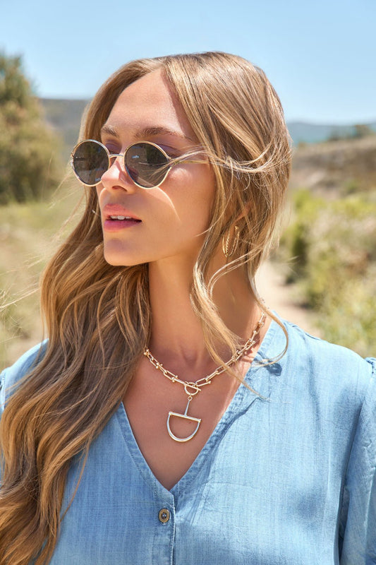 Woman with long blonde hair wears Gold Smoke Lens Round Sunglasses, a blue shirt, and a gold chain necklace outdoors on a sunny day.