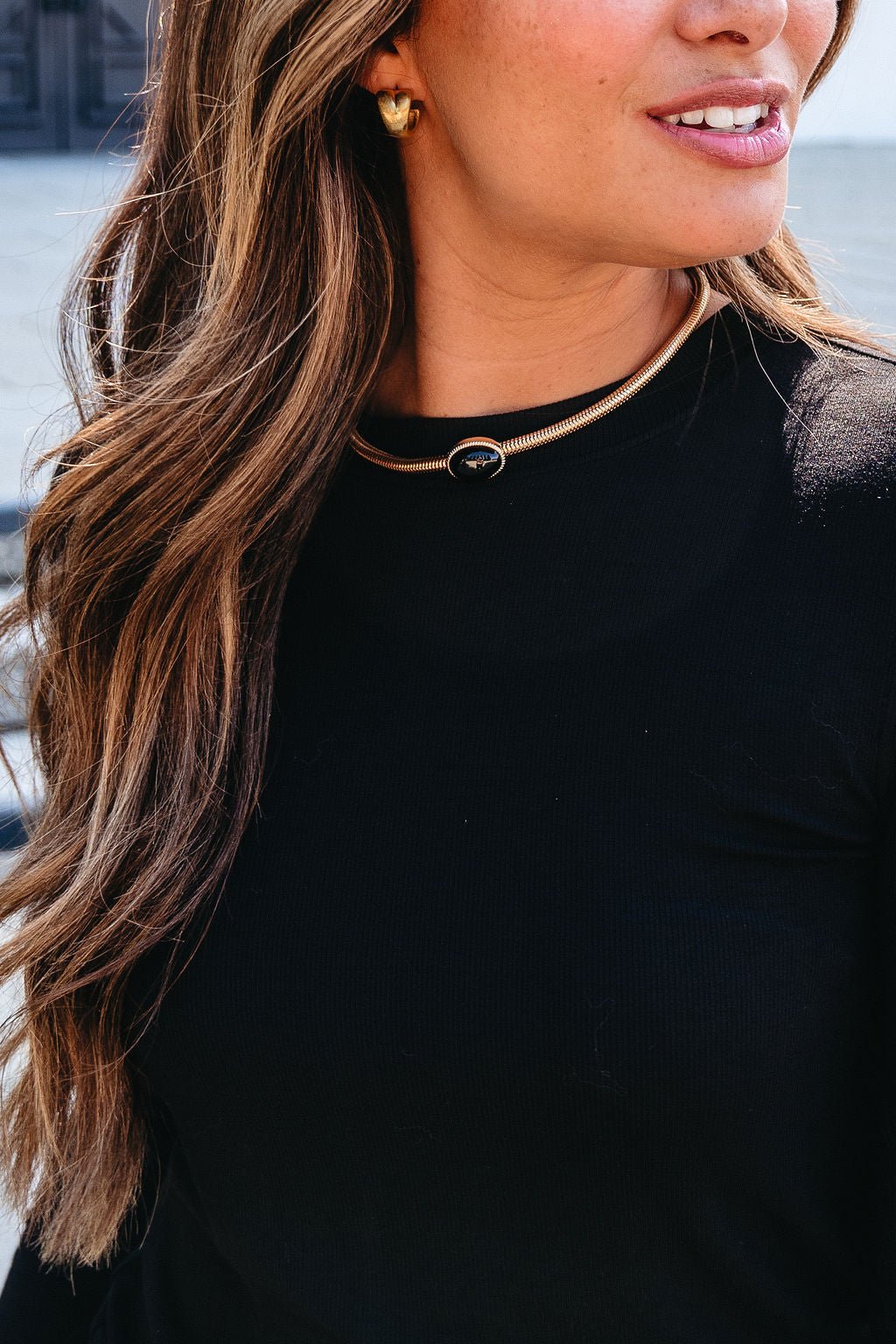 Woman with long wavy hair and black top wears Gold Snake Chain Black Oval Pendant Necklace and gold earrings, smiling slightly.