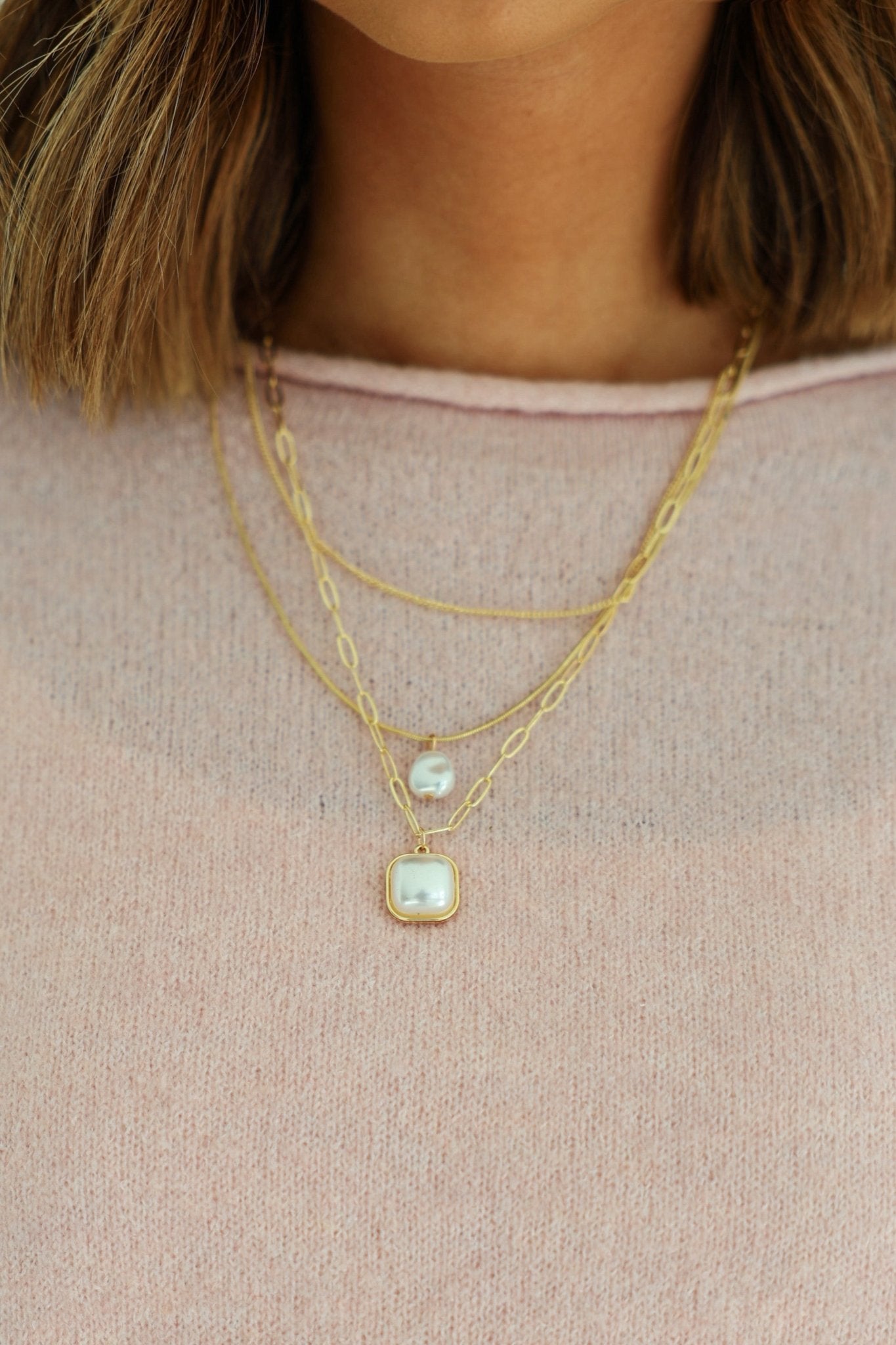 A woman wears a Gold Triple Layer Pearl Pendant Necklace over a light pink sweater.