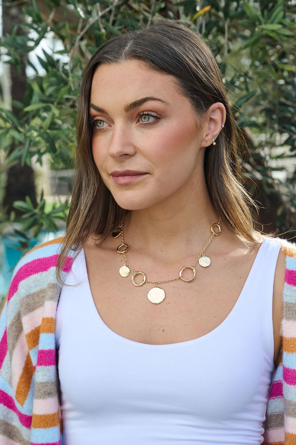 Woman with long brown hair wearing the Gold Warped Hoop & Coin Pendant Necklace, striped cardigan, and white top outdoors.