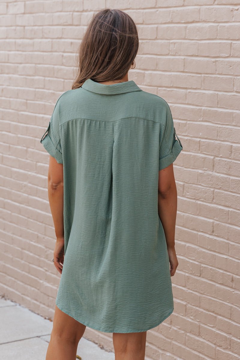 A woman with long brown hair models the Green Button Up Shirt Dress—FINAL SALE—beside a light brick wall.