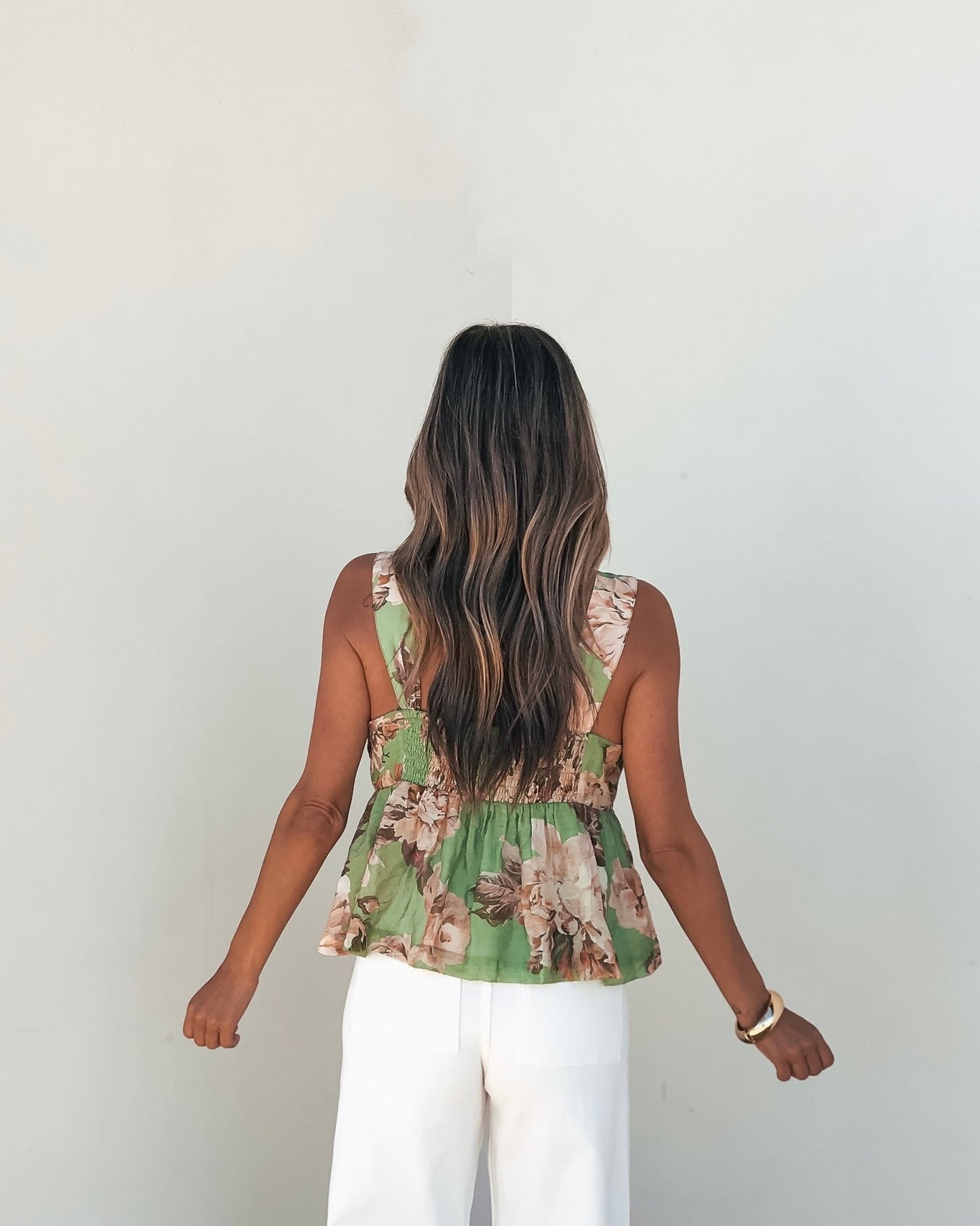 A woman with long wavy hair wears a Green Floral Silk Babydoll Tank Top and white pants, standing against a light wall.